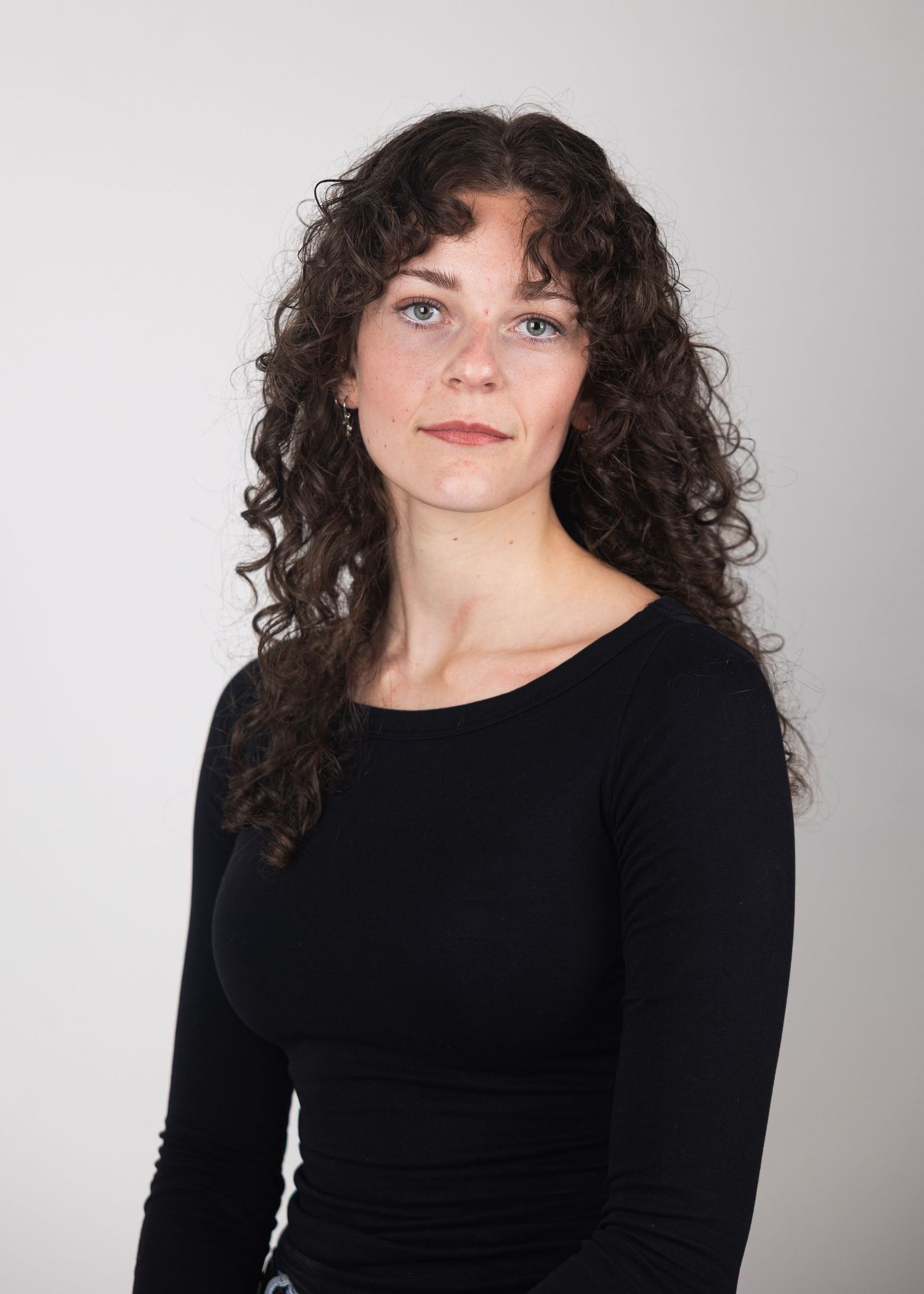 Young woman with long curly dark hair wearing a black long-sleeve top against a light background.