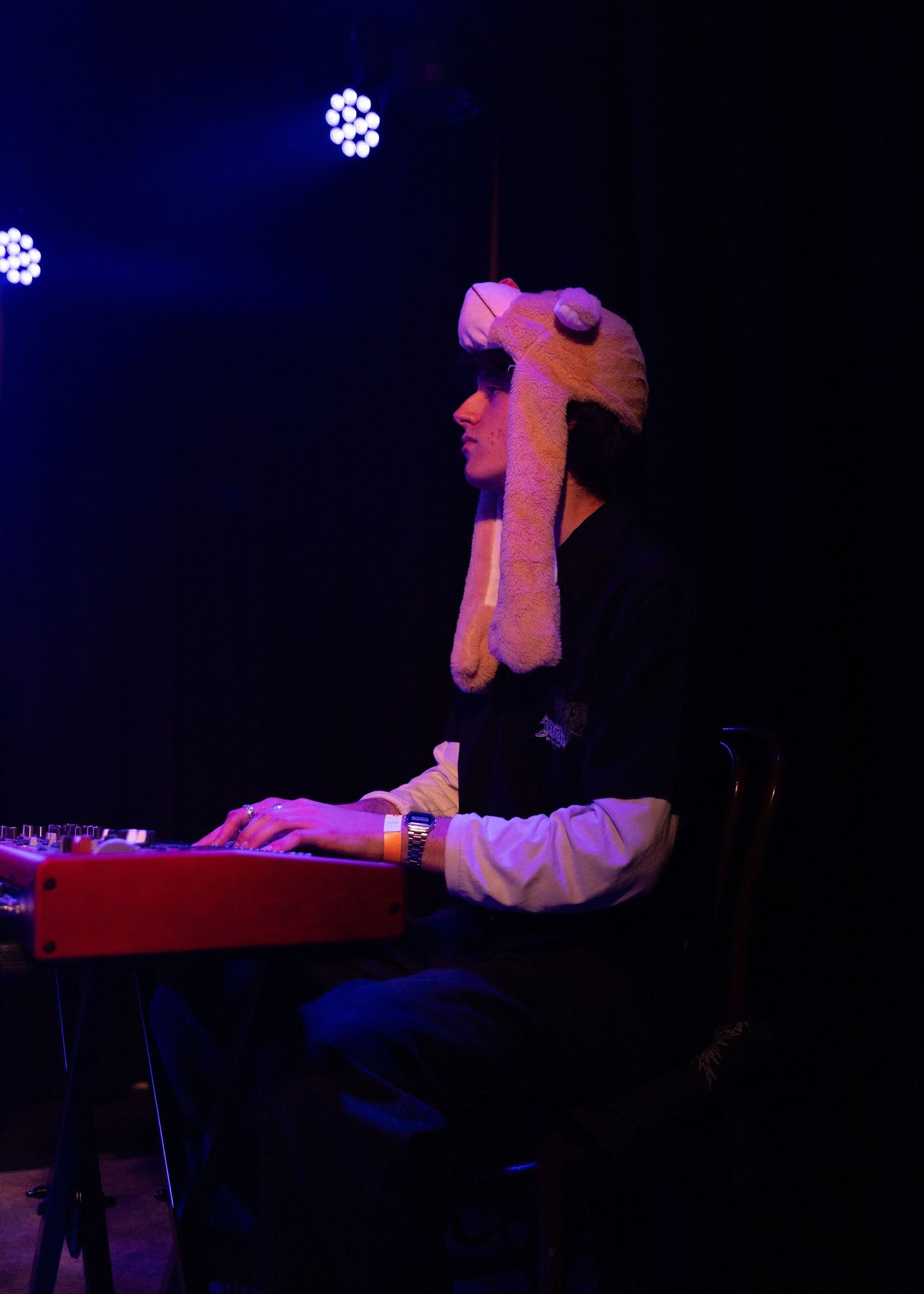 Musician wearing a novelty elephant hat plays a red keyboard on a dark stage with blue stage lighting.