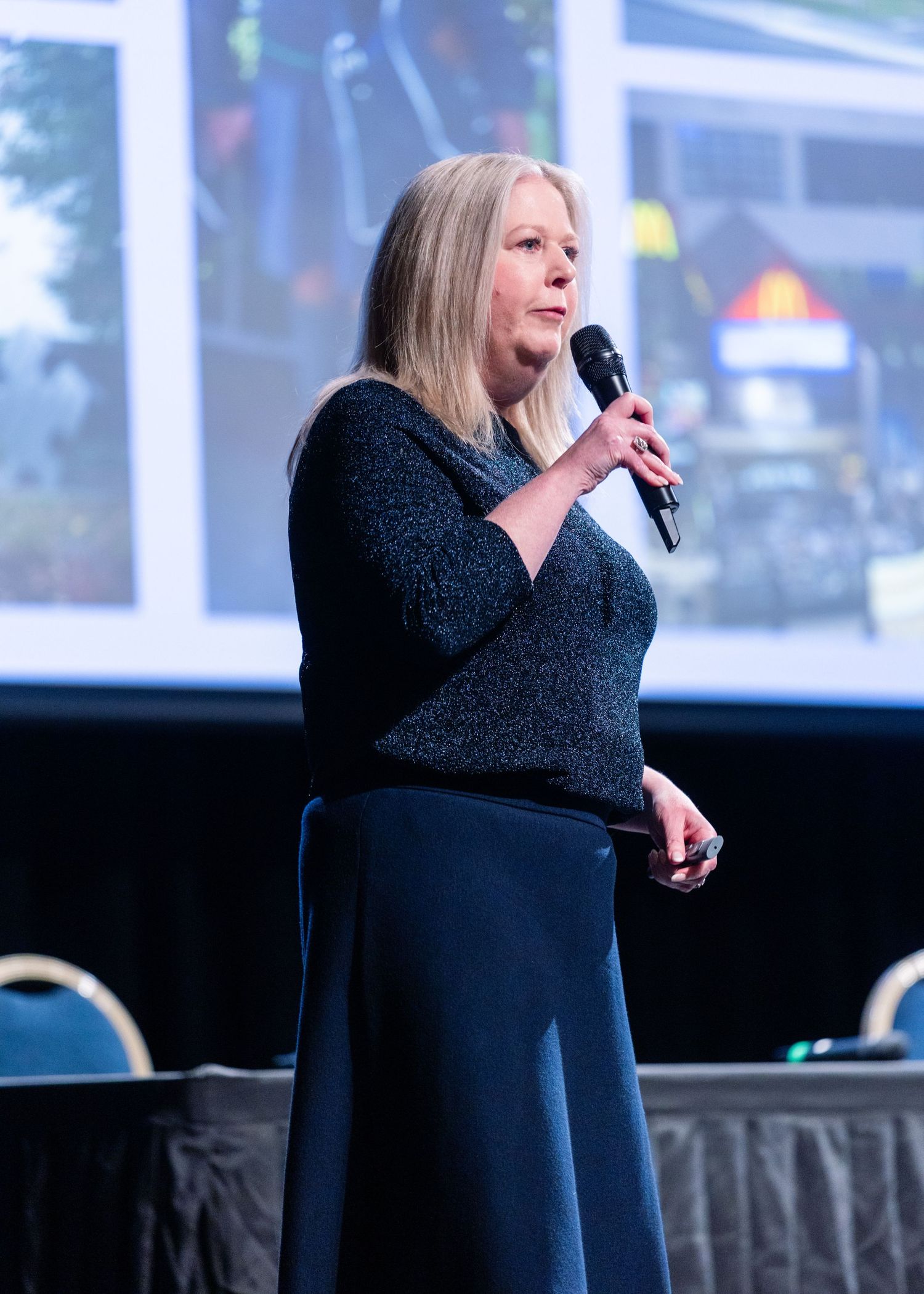A woman in a navy blue outfit speaks into a microphone at a business conference presentation.