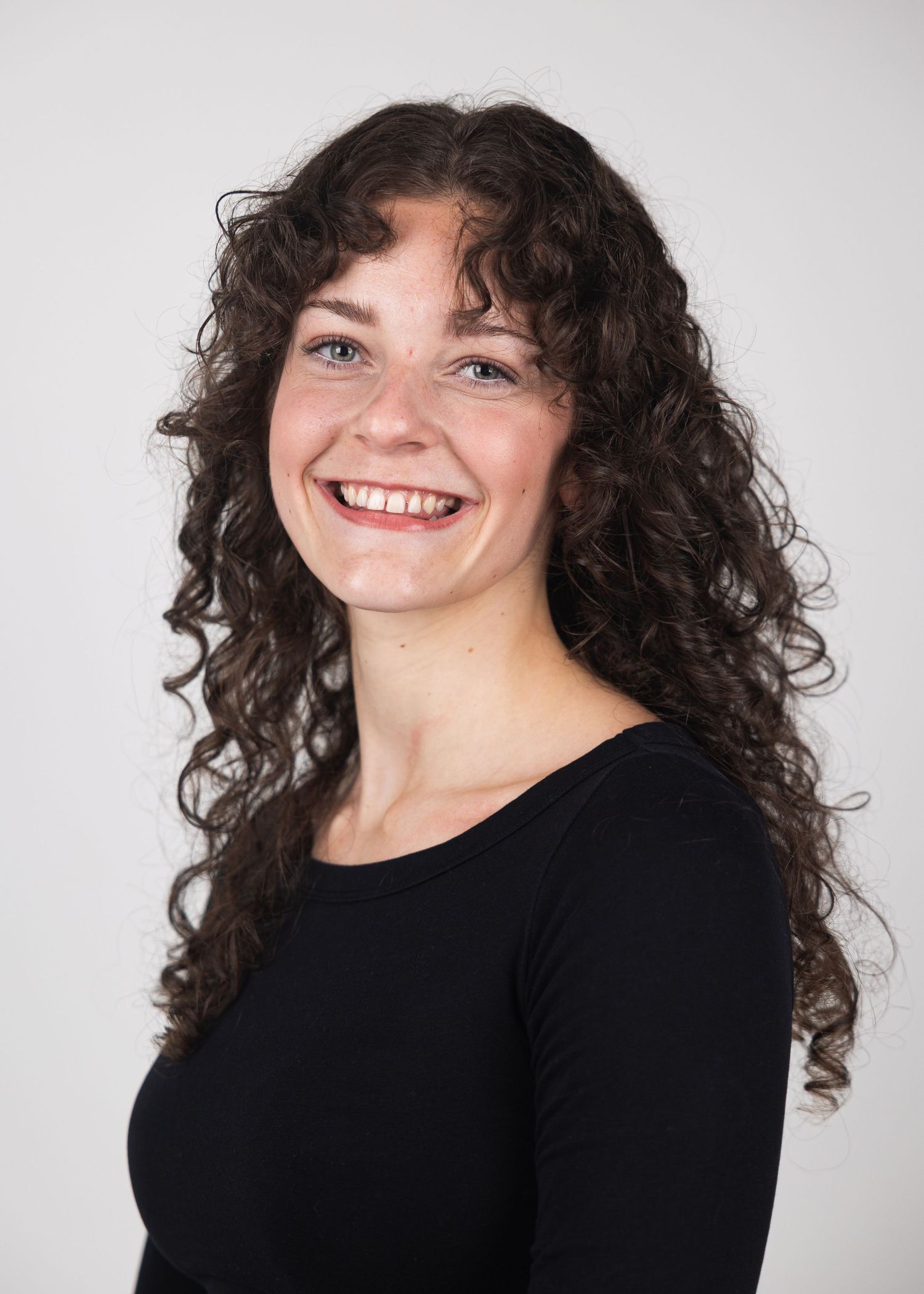 Young woman with curly brown hair smiling confidently in a black top against a white background.