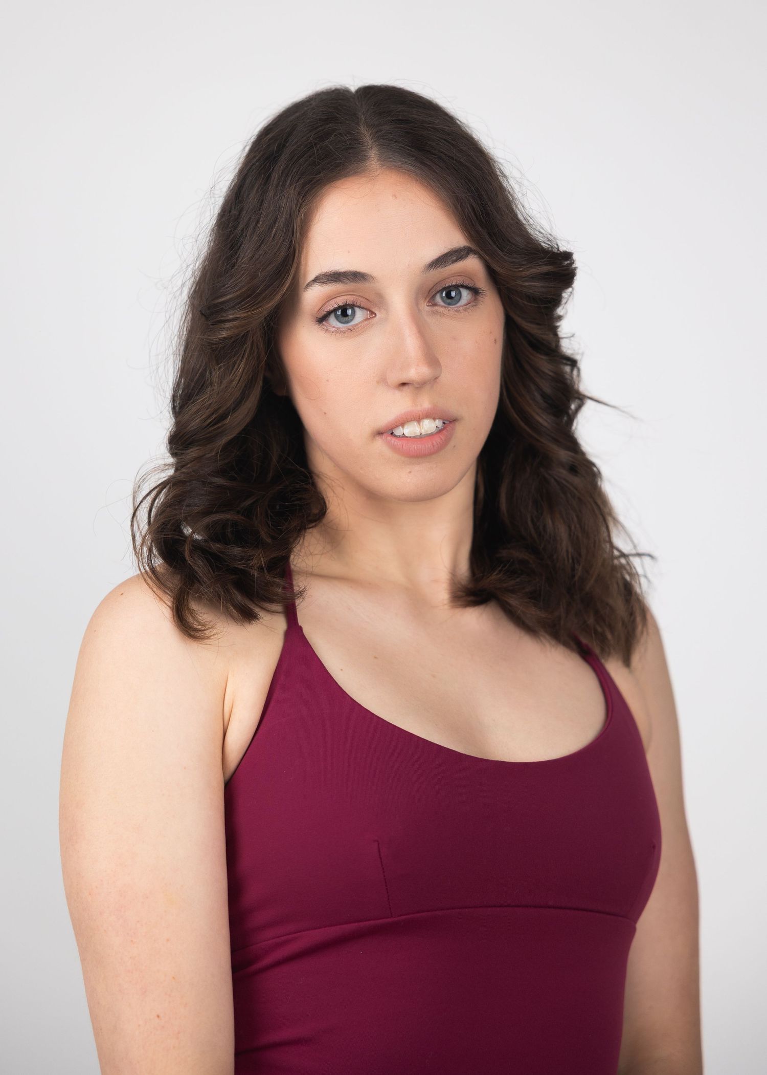 Young woman with dark wavy hair and blue eyes wearing a burgundy tank top against a light gray background.