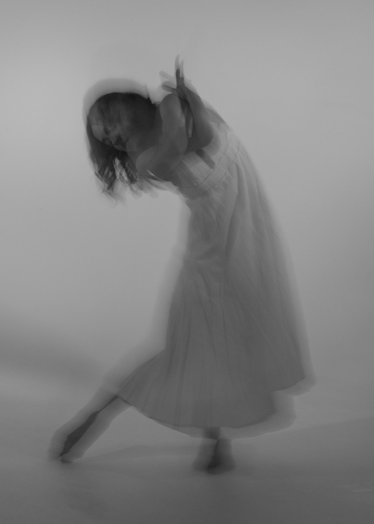 Blurred black and white photo of a dancer in a white dress, gracefully bending with arms raised.