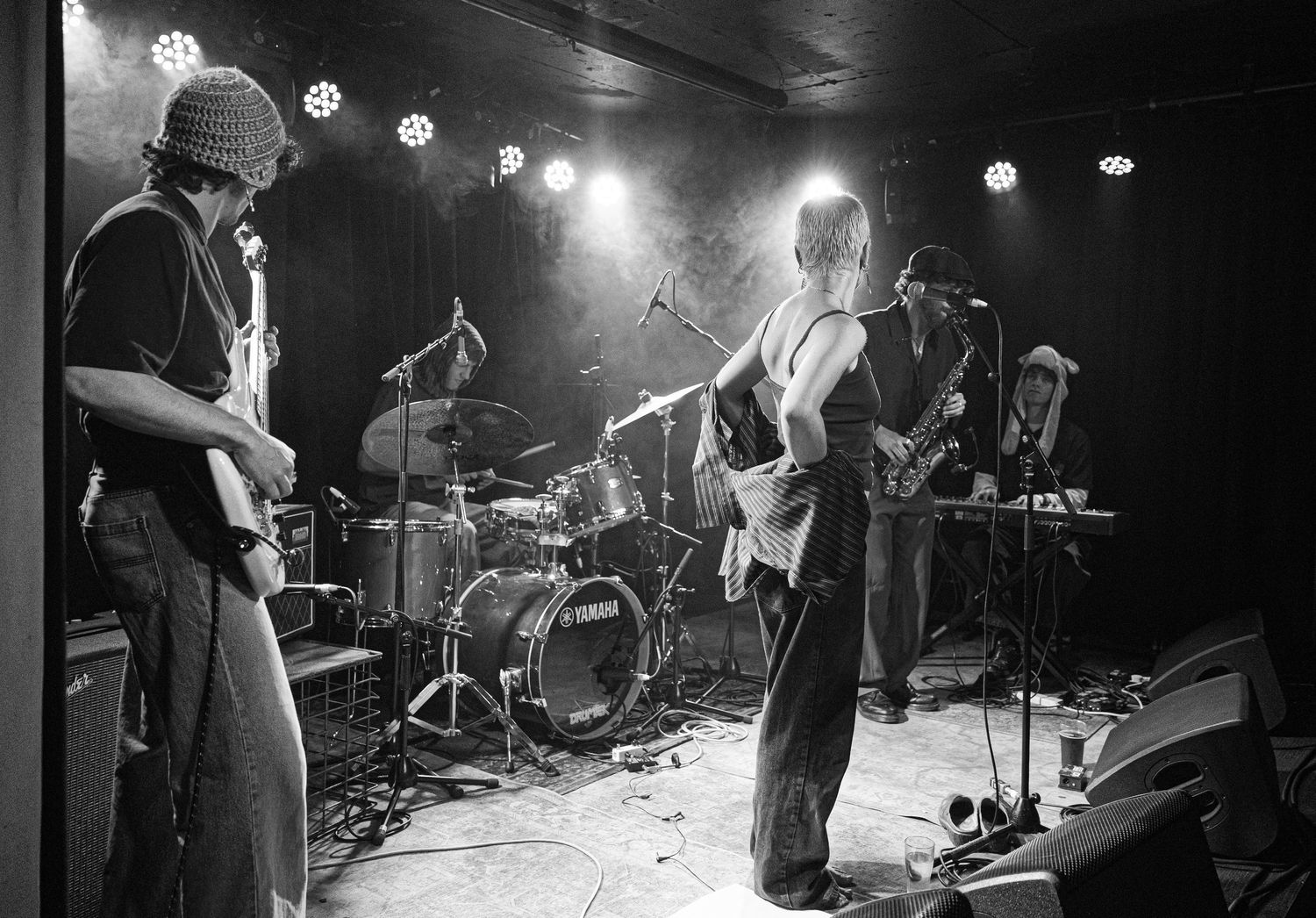 Black and white photo of a rock band performing on stage with guitars and drums under bright stage lights.