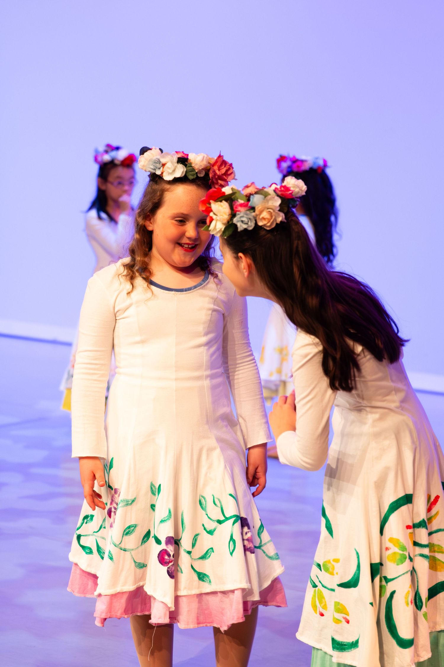 Girls in white floral dresses and flower crowns perform on stage during a children's dance recital.