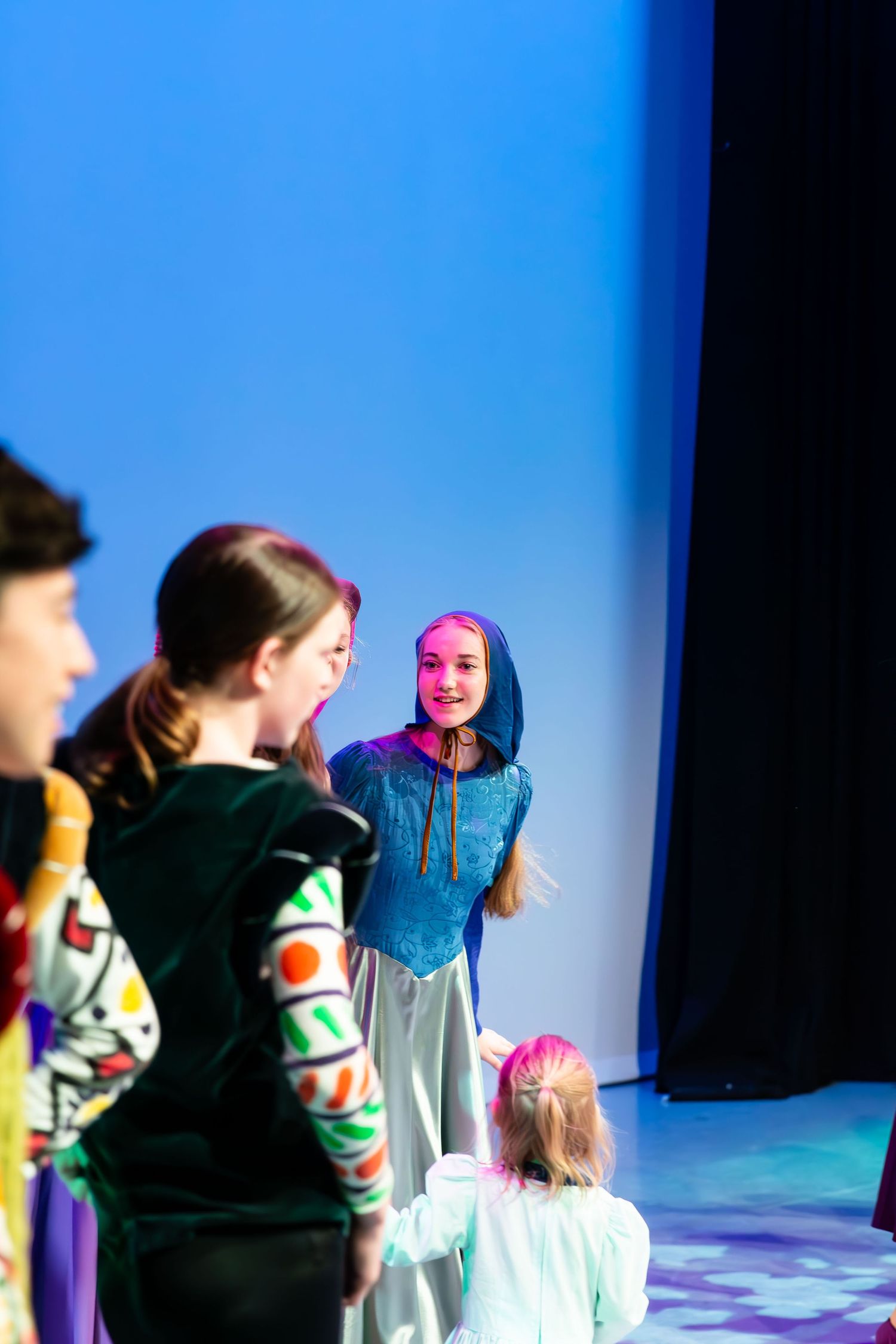 Children in colorful costumes perform on a blue-lit stage during a school theater production.