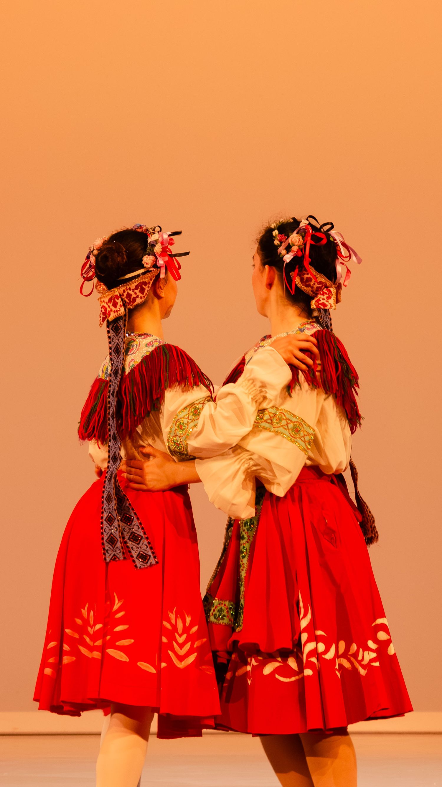 Traditional dancers in red and white costumes perform a graceful duet on stage against an orange backdrop.