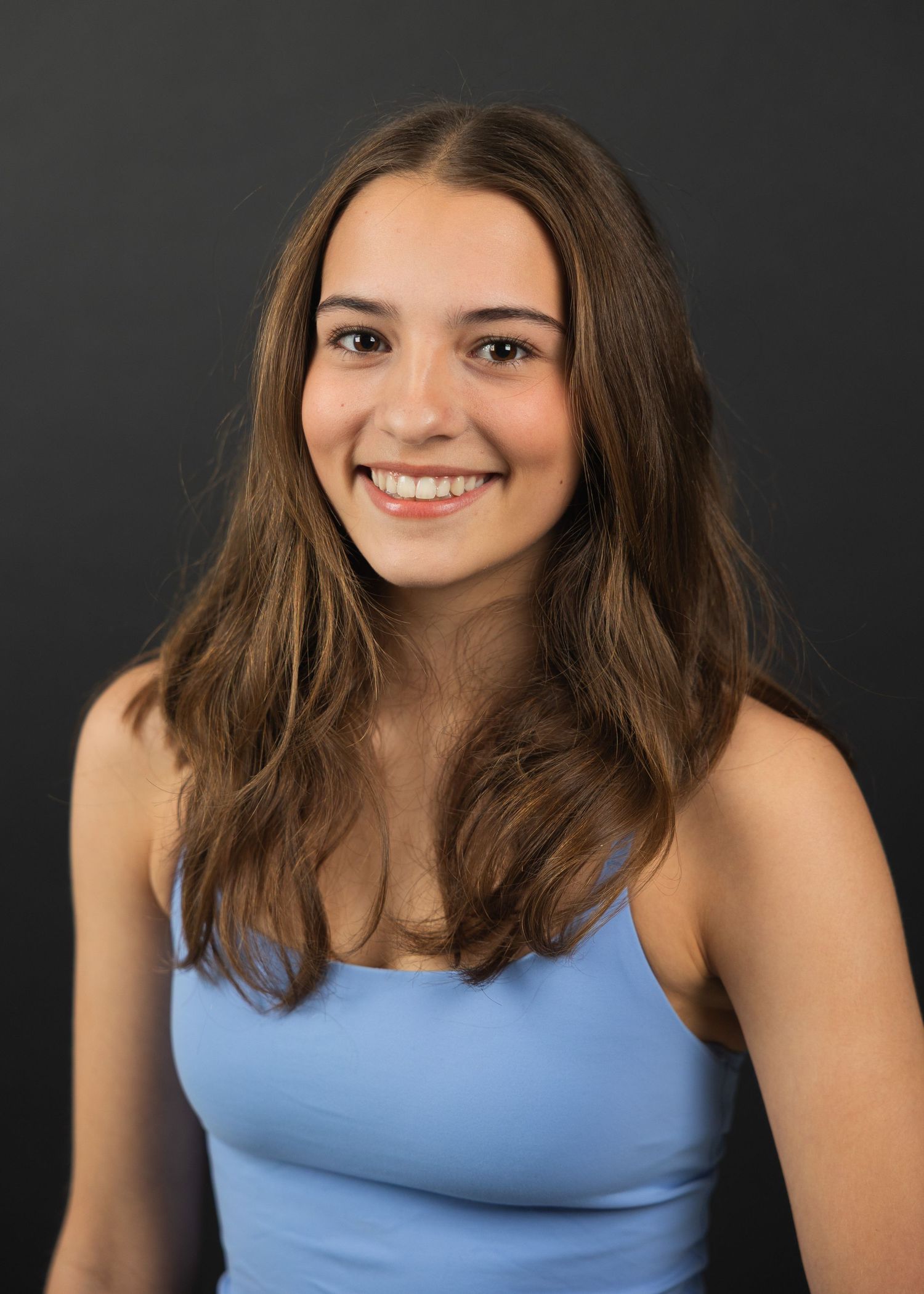 Young woman with long brown hair smiling, wearing a light blue top against a dark background.