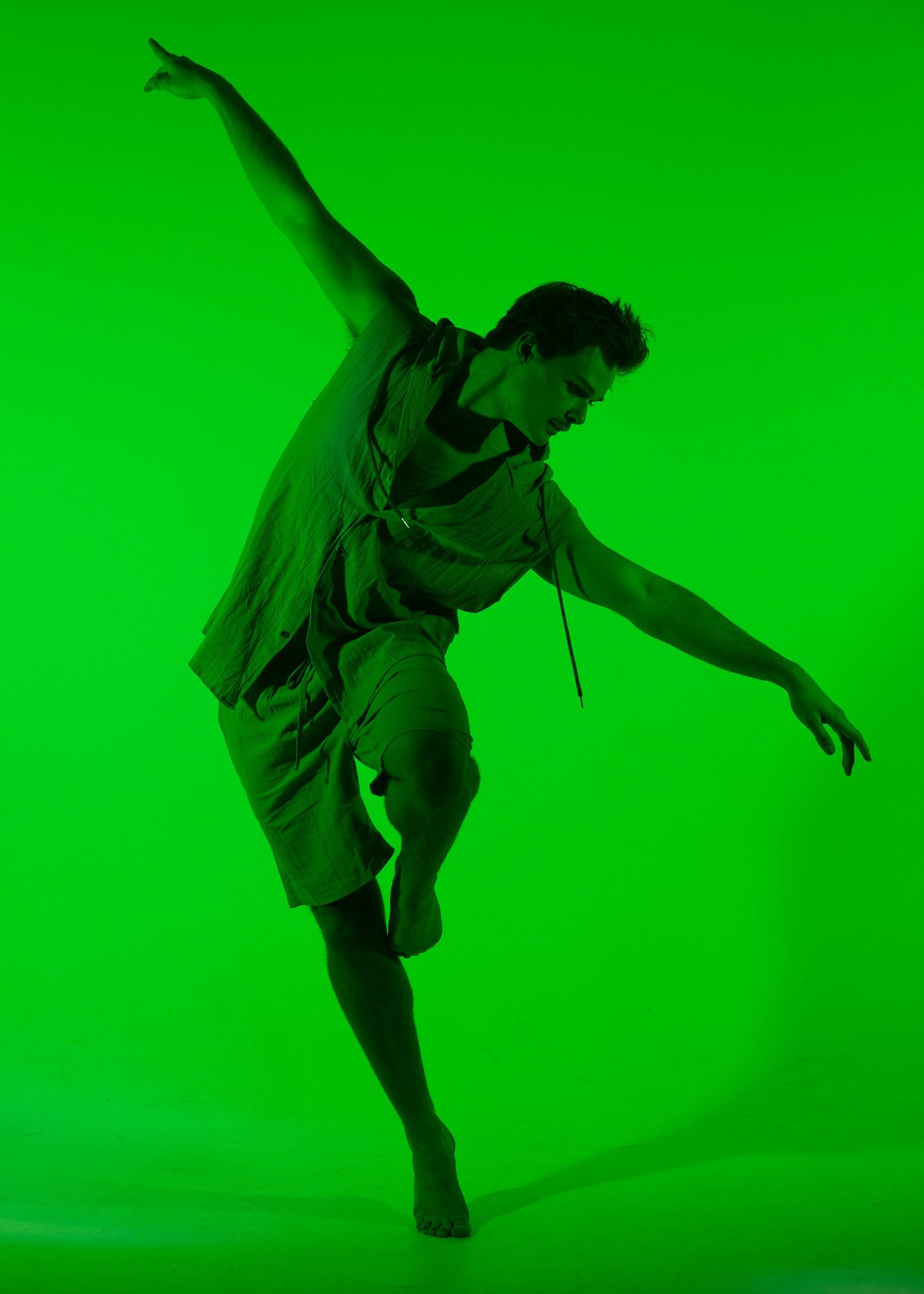 A male dancer leaps dynamically against a vibrant green background, wearing shorts and a loose top.