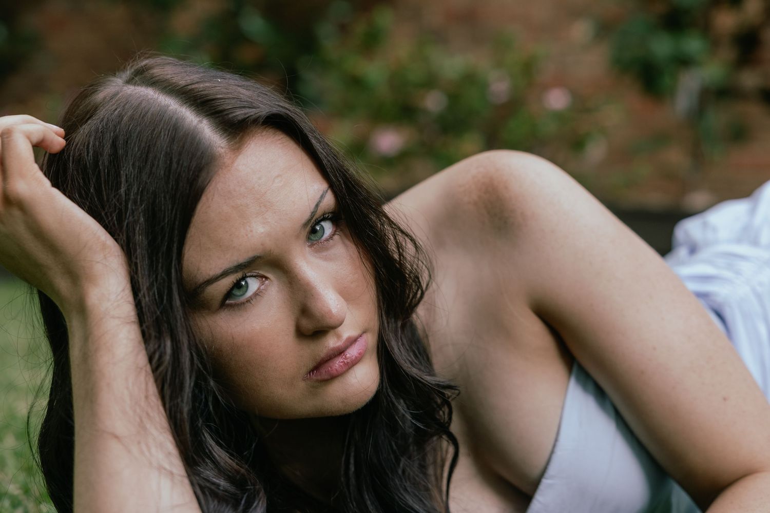 Young woman with dark wavy hair lying on grass, gazing at camera with a contemplative expression outdoors.