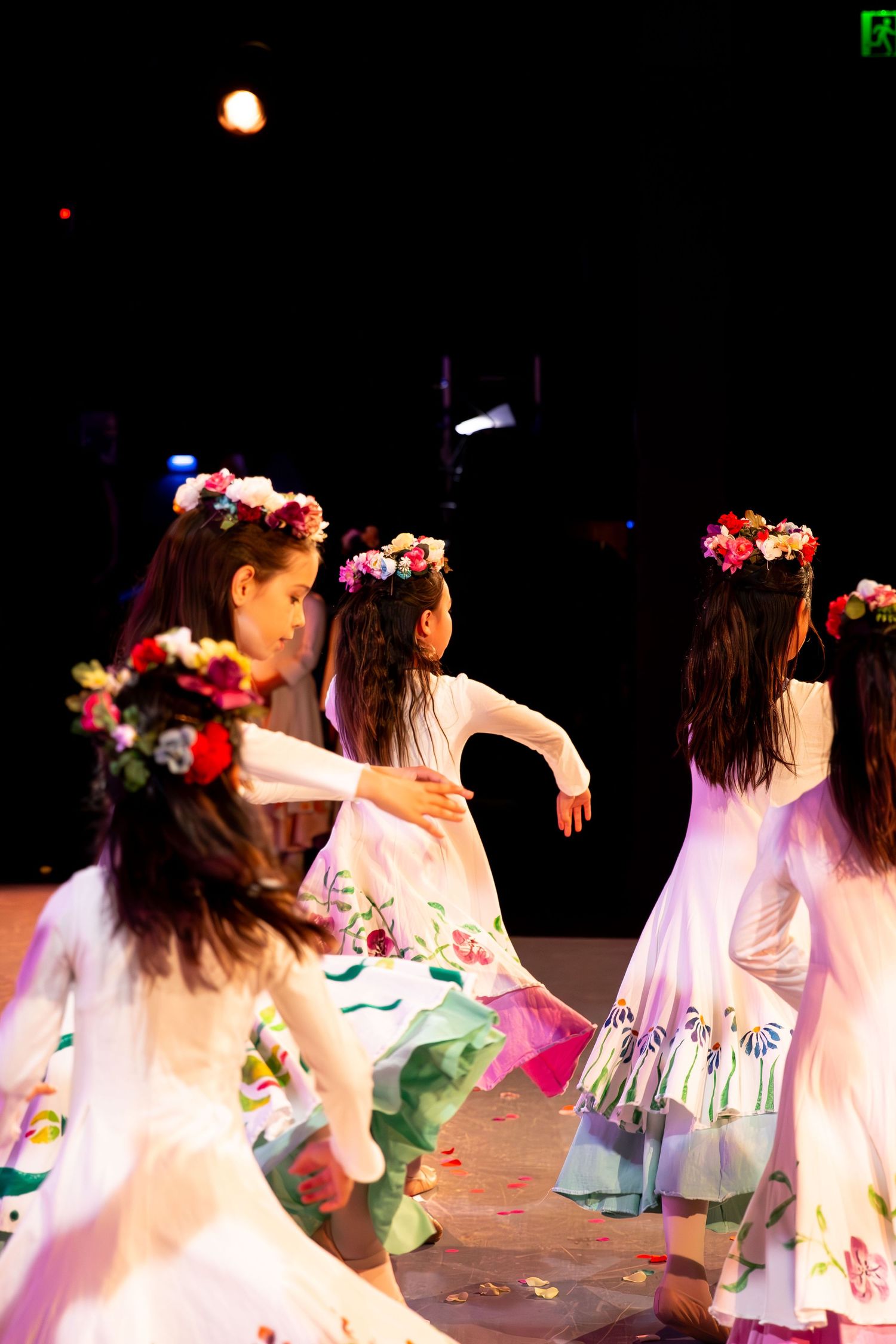 Young girls in floral crowns and white dresses perform a traditional folk dance on a dimly lit stage.