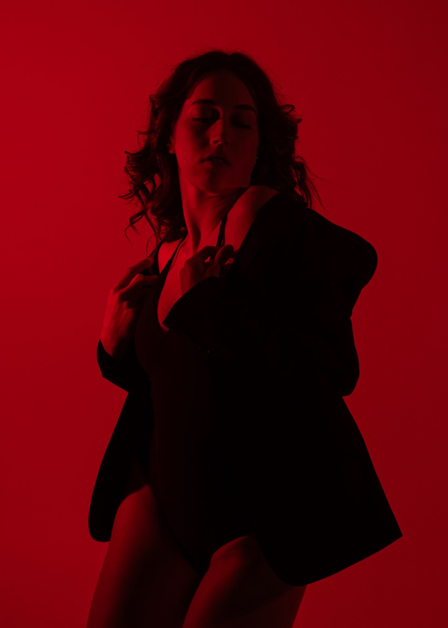 A woman in a black blazer poses dramatically against a vibrant red background in moody, artistic boudoir photography.