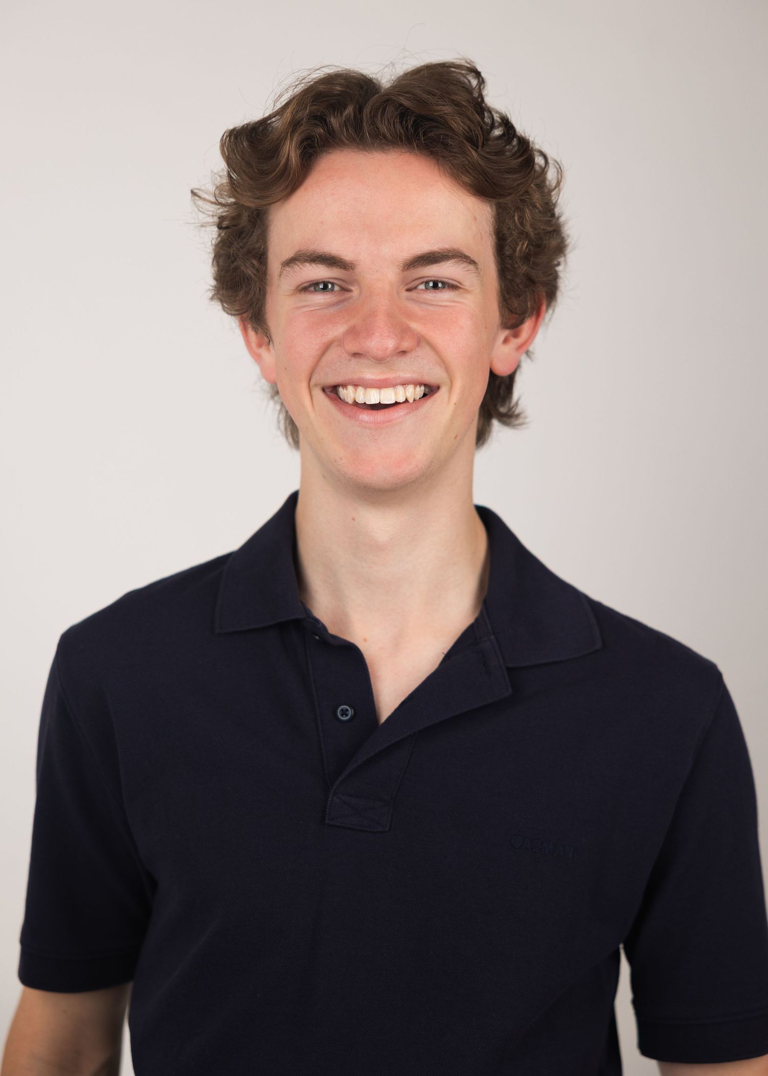 Smiling young man with curly brown hair wearing a navy polo shirt against a light gray background.