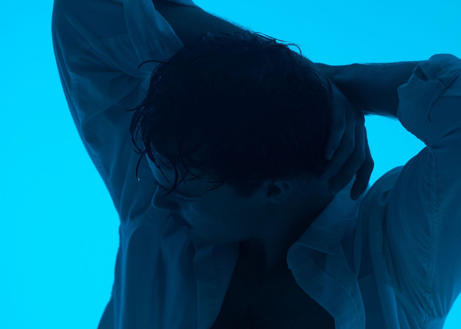Person with hands behind head posed against vibrant cyan blue background in artistic moody portrait photography.