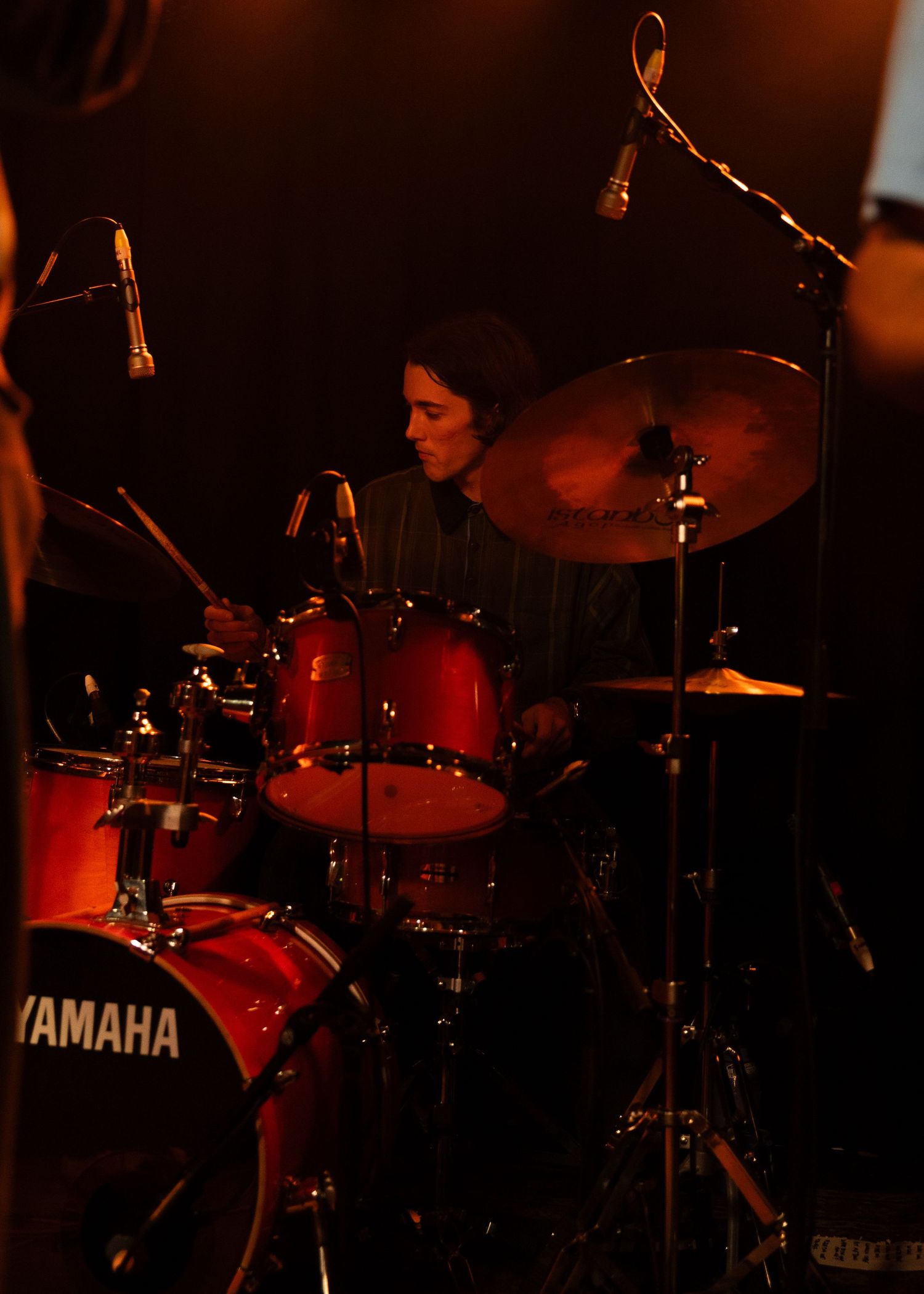 Drummer playing red Yamaha drum kit with cymbals on a dimly lit stage during a live performance.