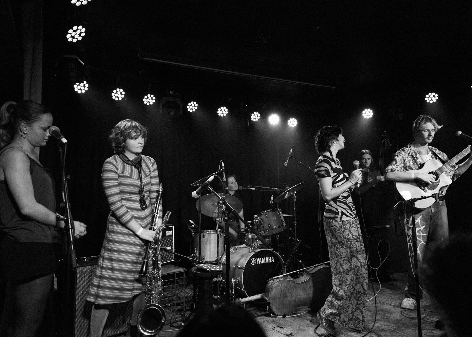Black and white photo of a live band performing on stage with dramatic lighting and multiple musicians playing instruments.