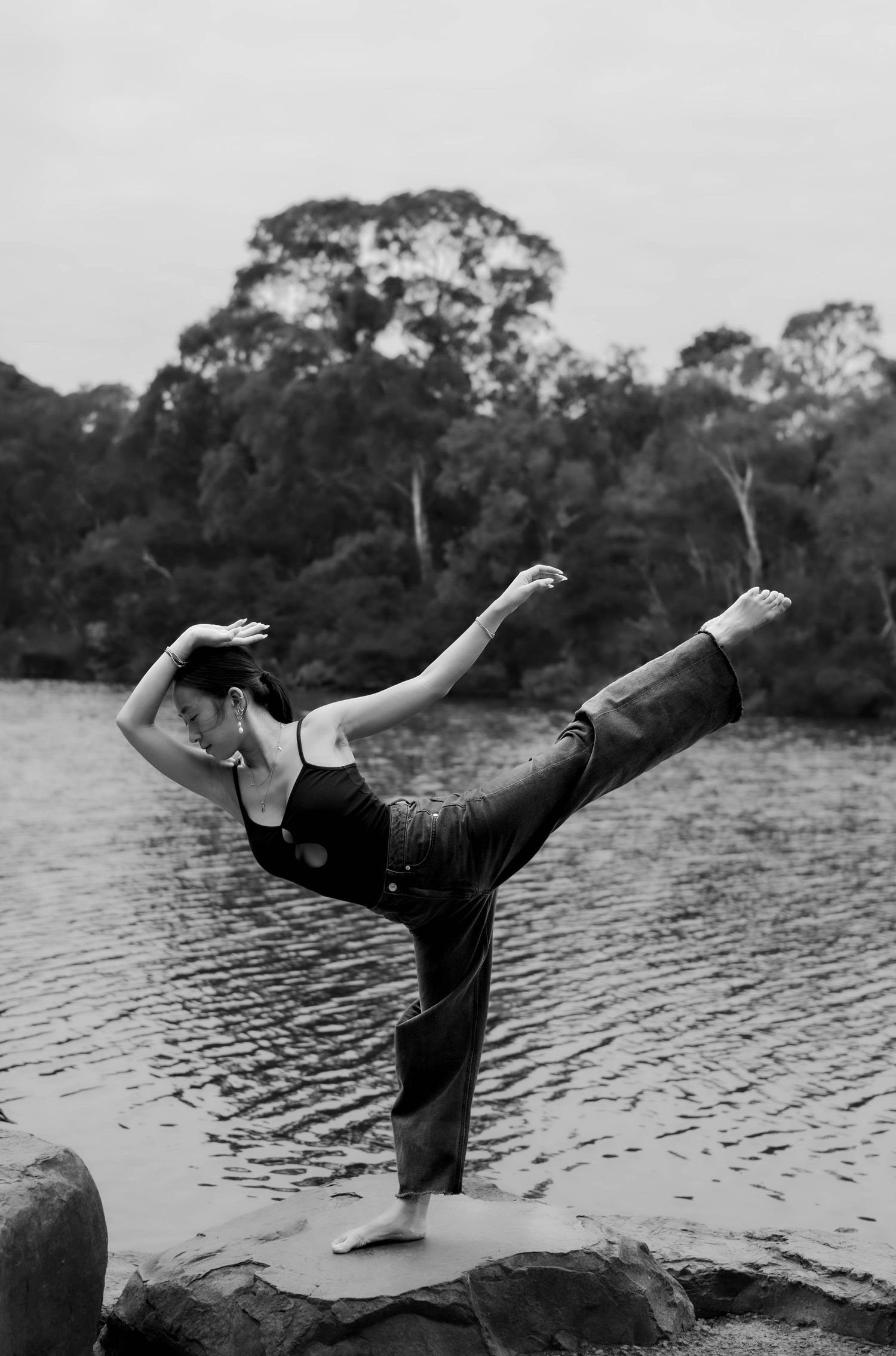 Black and white photo of a woman balancing on one leg in a yoga pose on rocks by a tranquil river surrounded by trees.