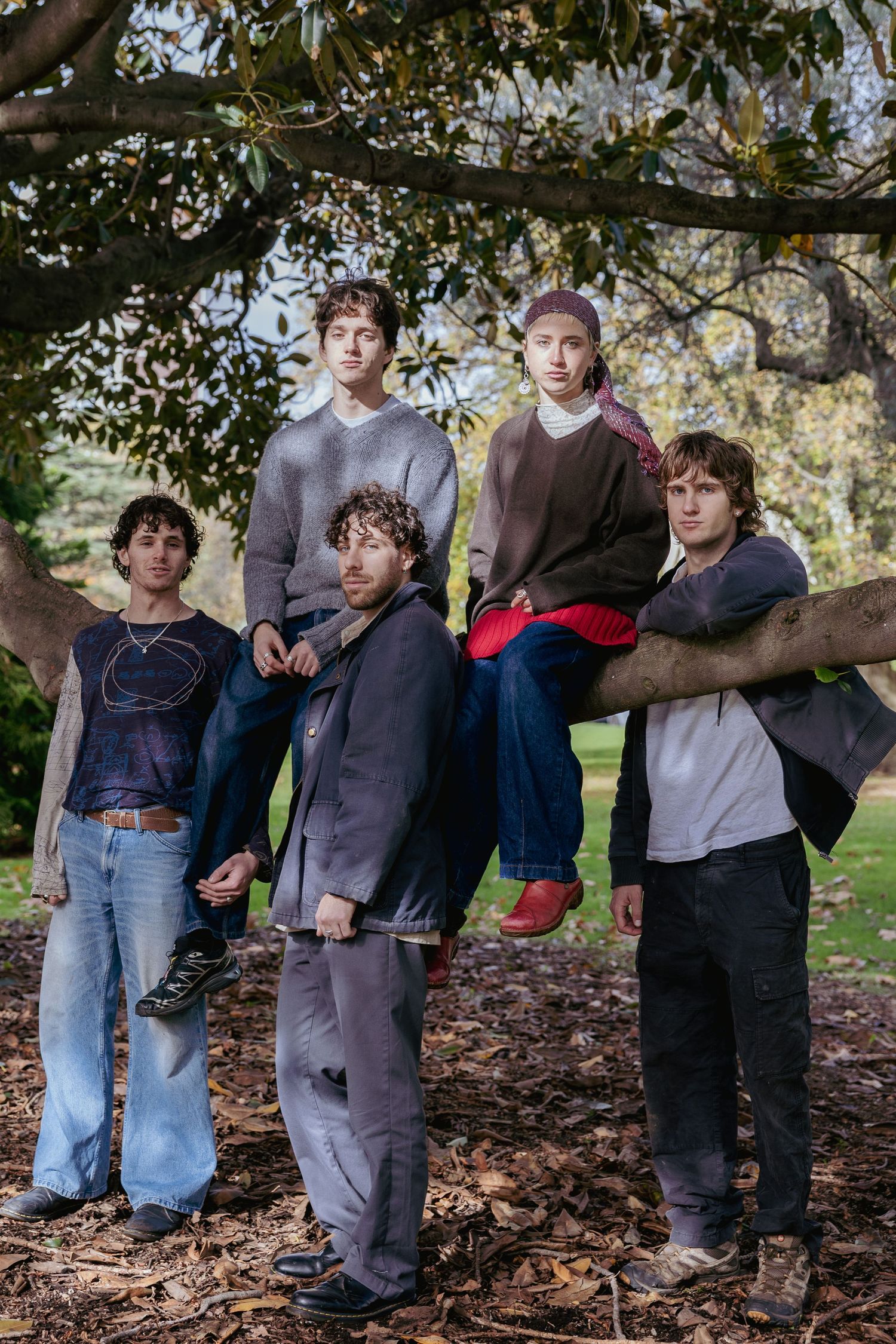 Five young people pose among tree branches and fallen leaves in a park, with casual indie style clothing.