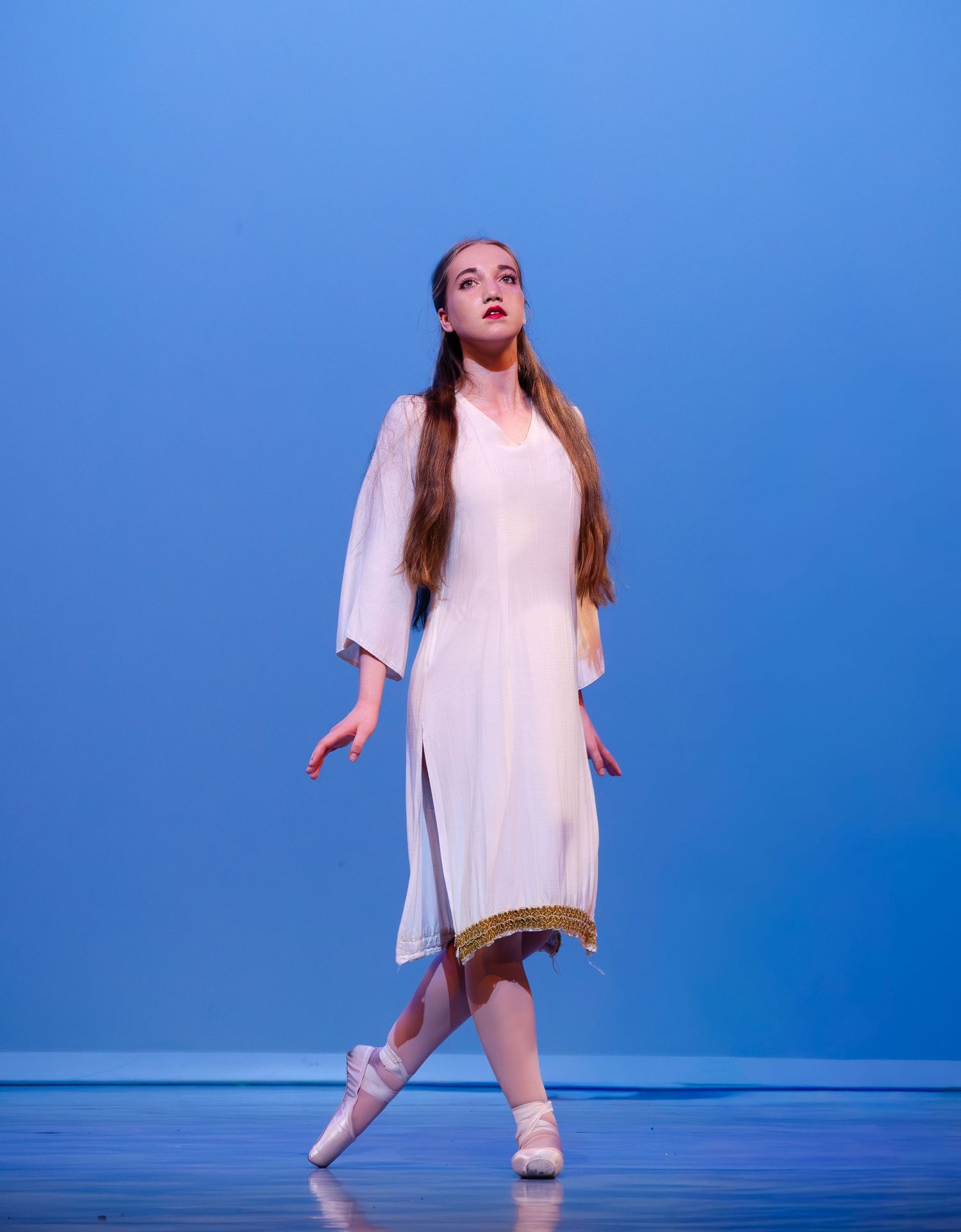 Ballet dancer in white dress performs en pointe against blue backdrop during stage performance.