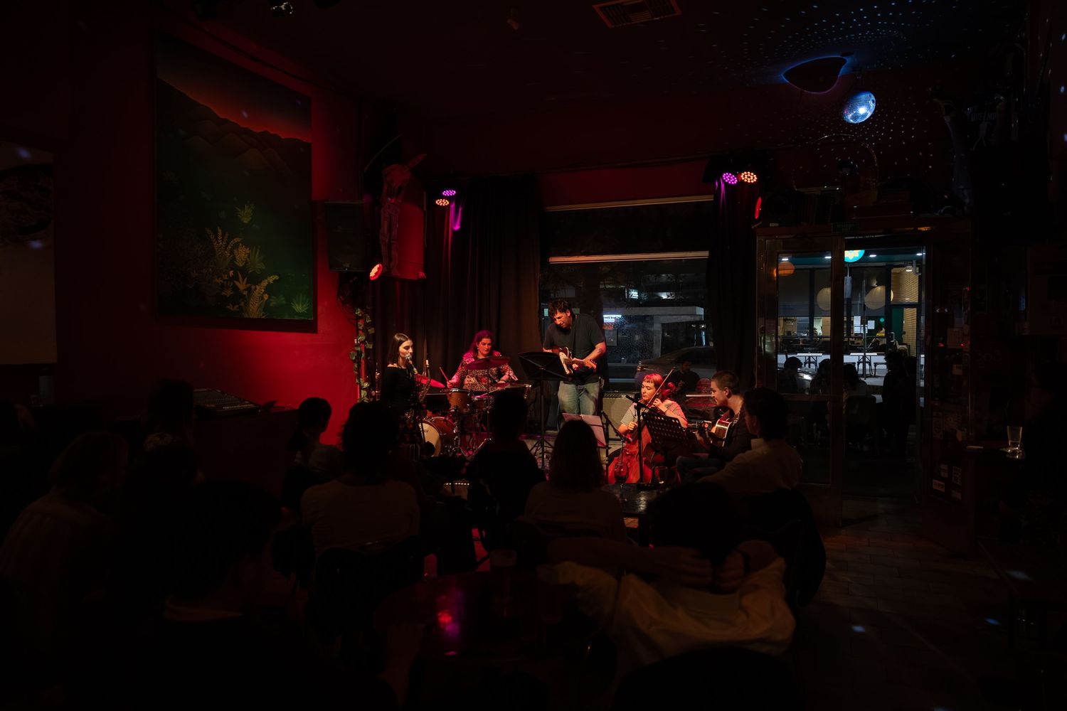 A dimly lit music venue with a band performing on stage under red lighting and audience members watching in the darkness.