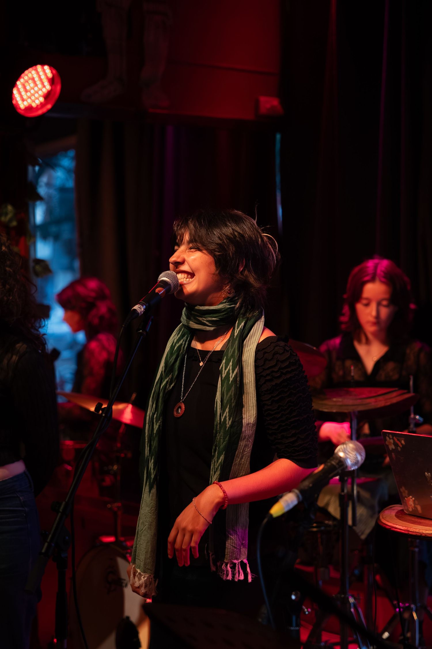 A performer sings into a microphone on a dimly lit stage with red and blue lighting while wearing a scarf.