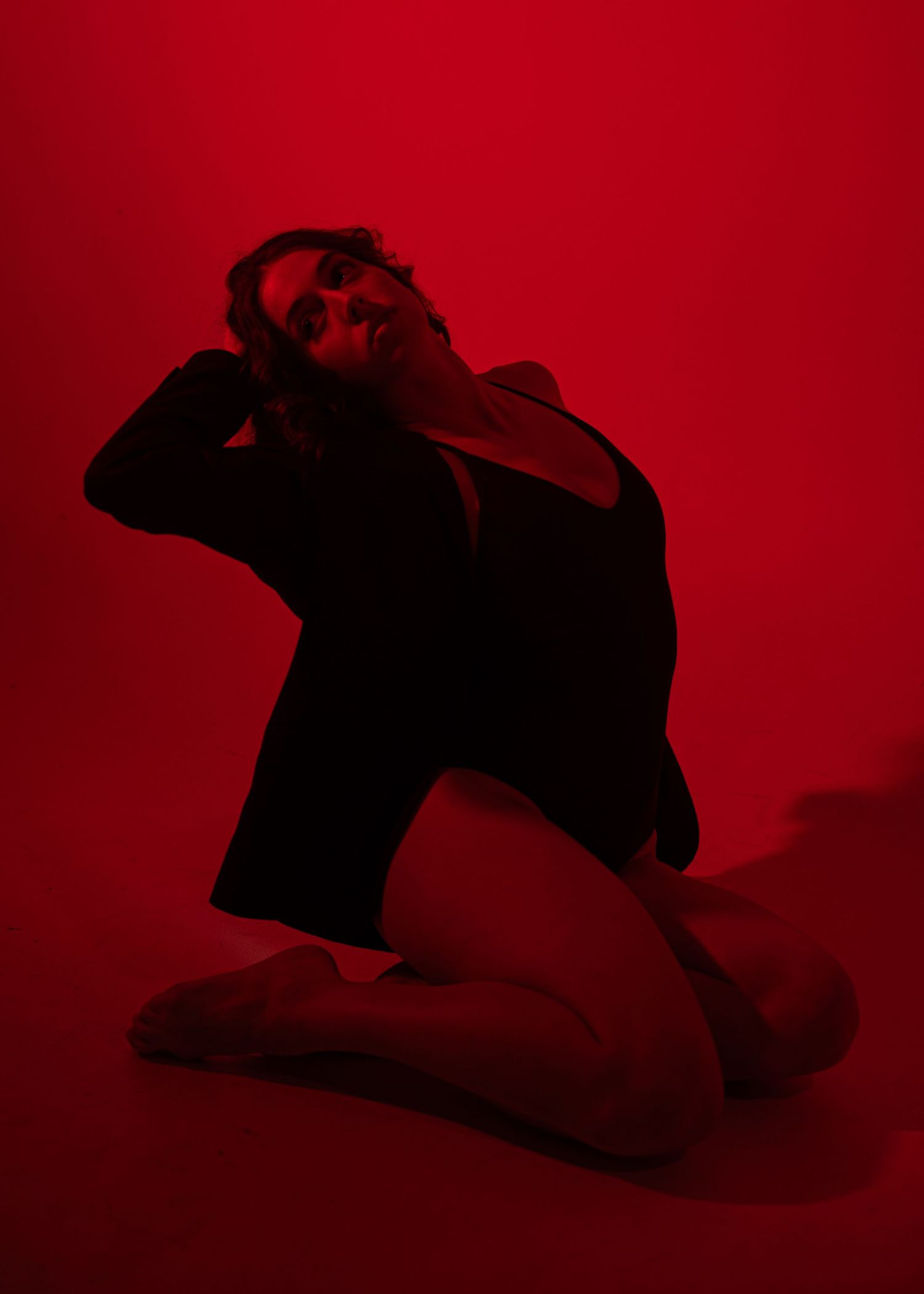A woman in a black outfit kneels on the floor against a red background, head tilted back in an expressive pose.