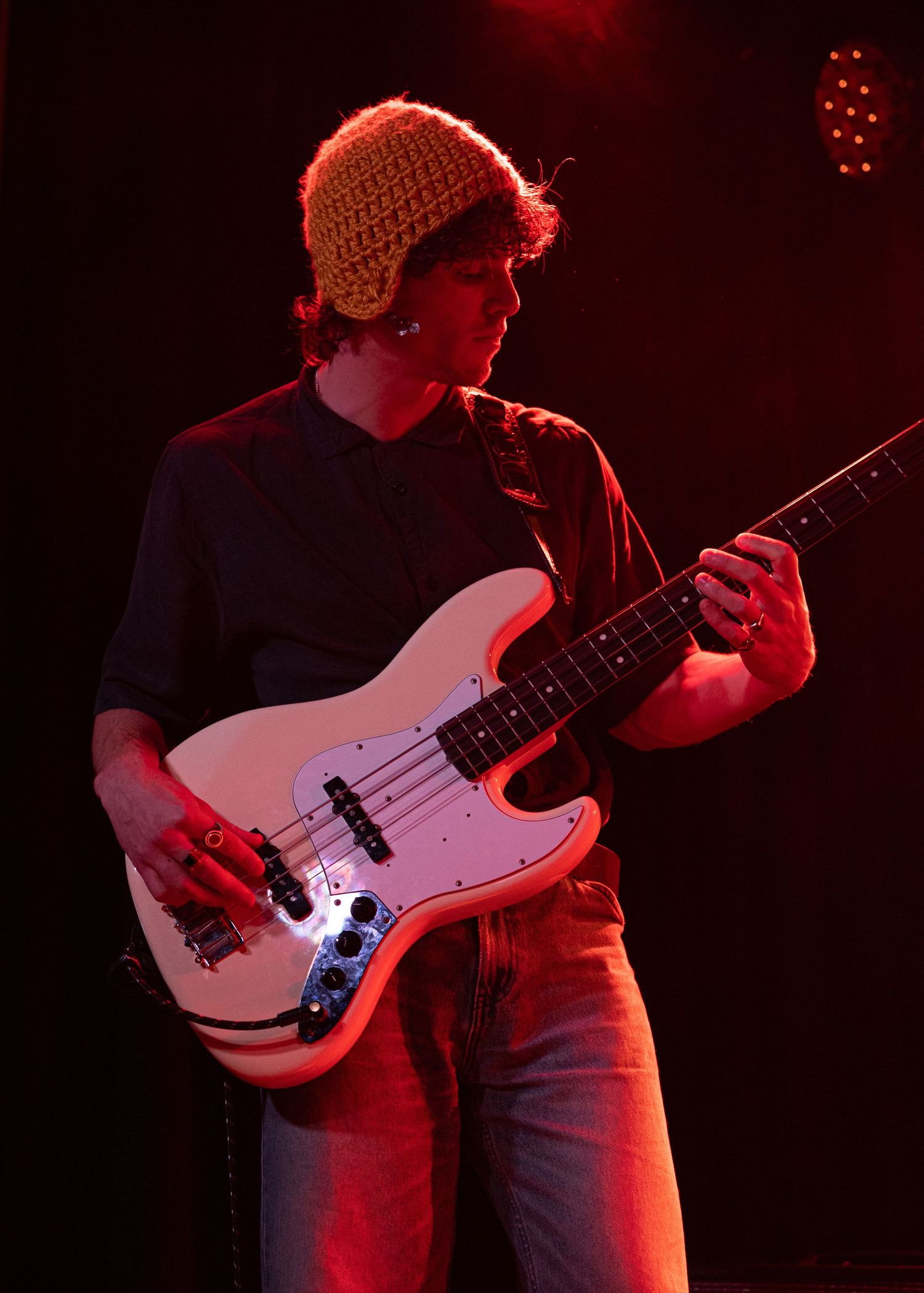 Close-up of male bassist in knit hat playing white Fender bass guitar on red-lit stage.