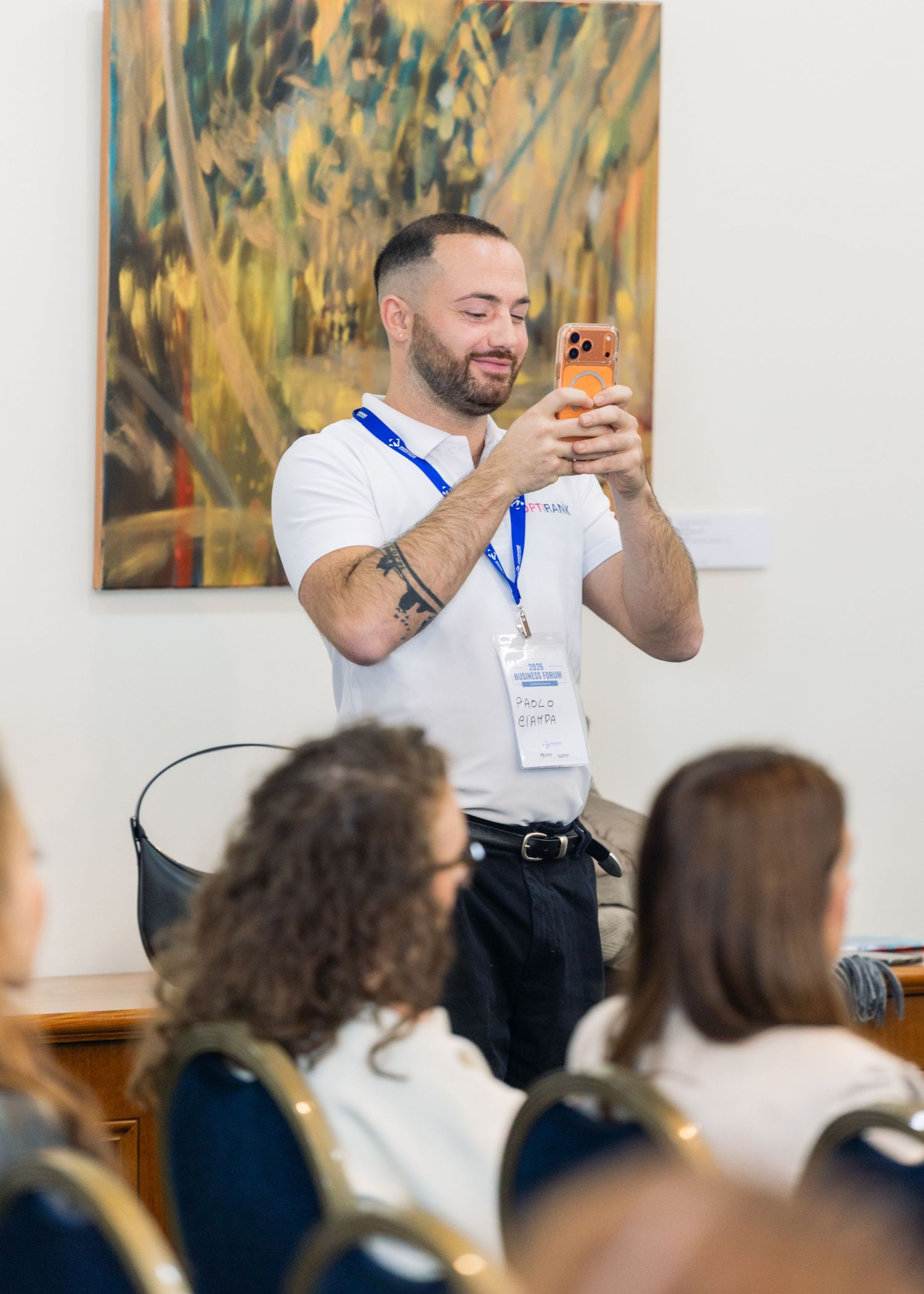 A man in a white polo shirt and lanyard smiles while taking a photo on his phone at an indoor conference event.