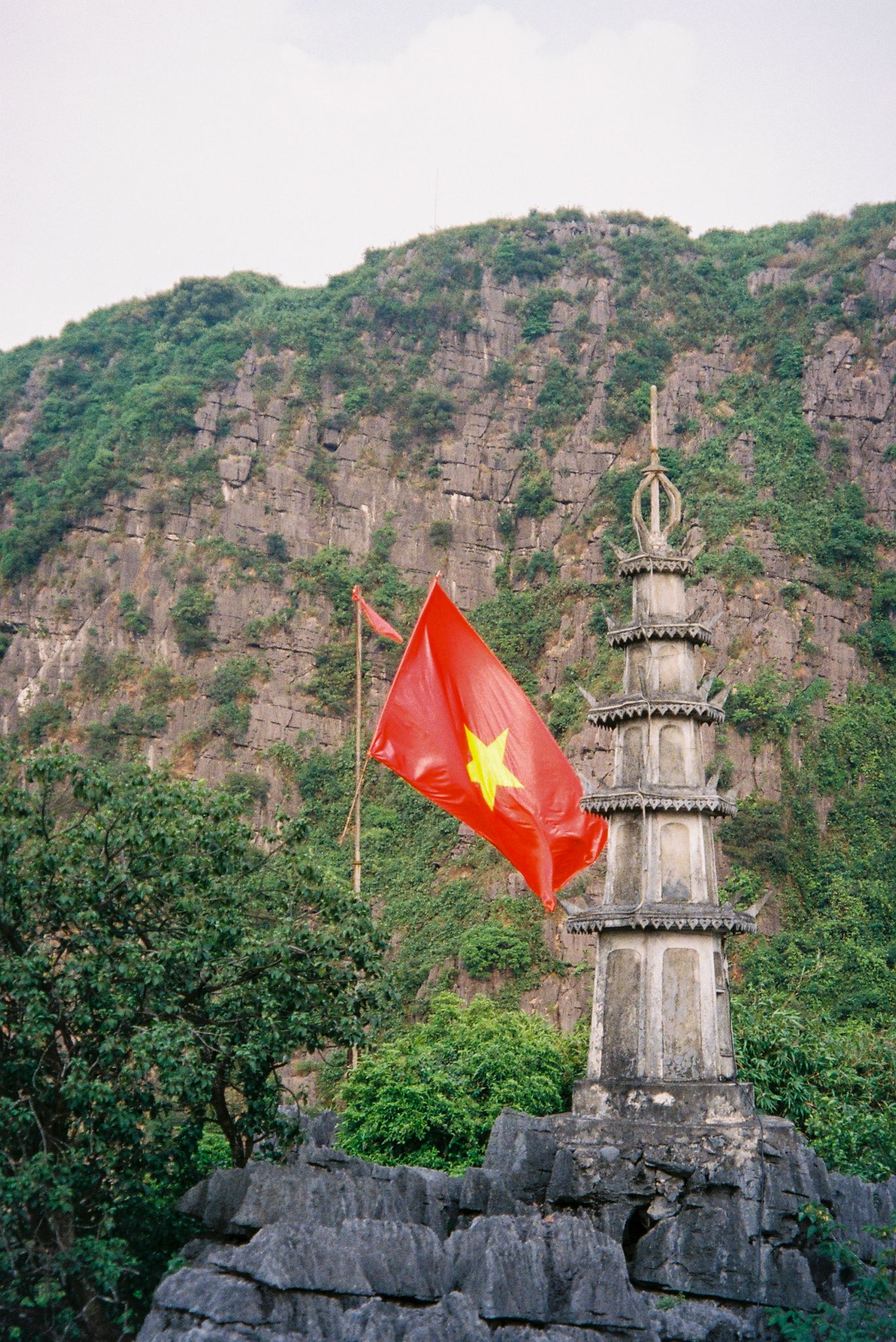 Vietnamese flag waves atop ancient stone watchtower against dramatic mountain cliffs and lush green forest backdrop.