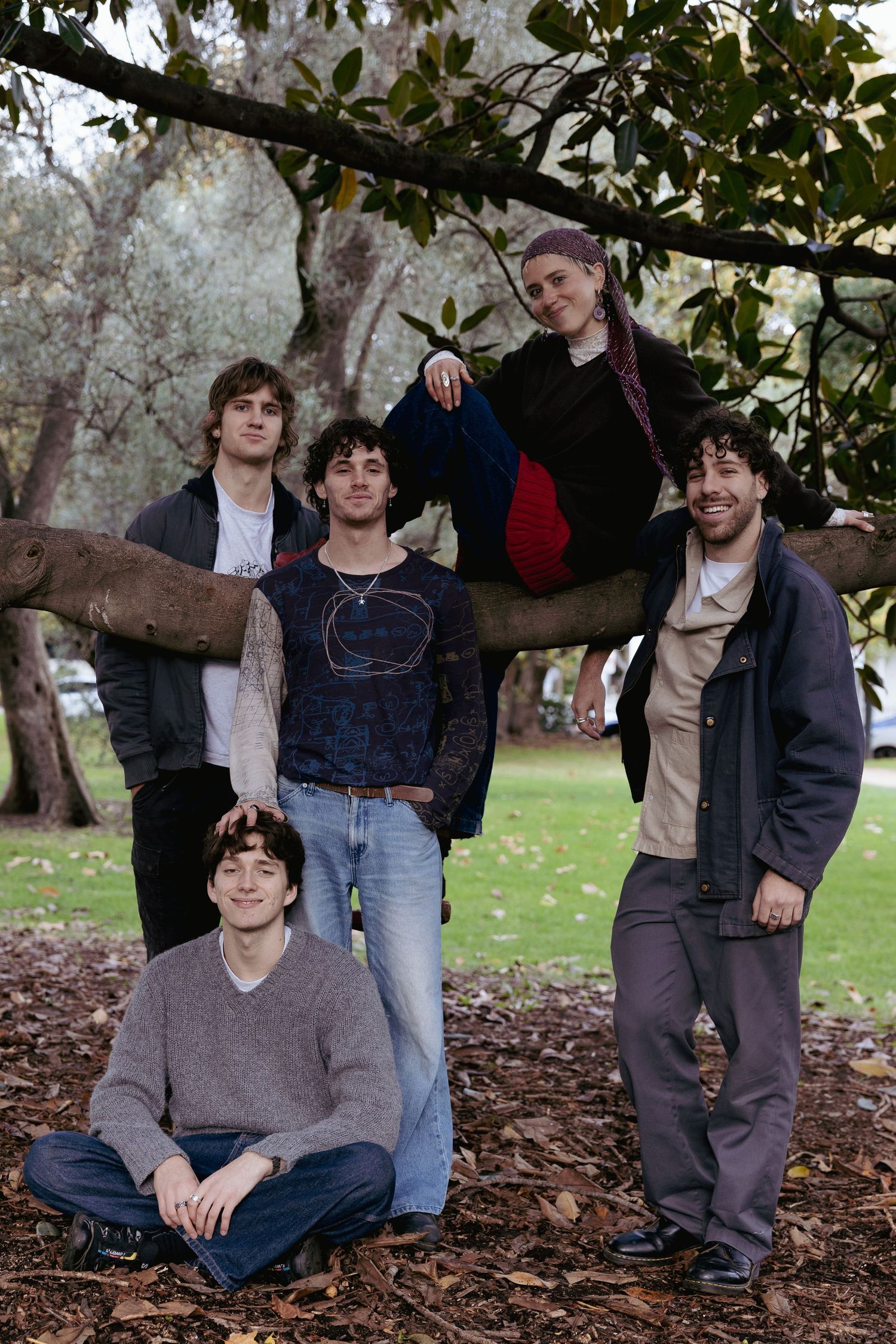 Five young people pose among tree branches and autumn leaves in a park setting.