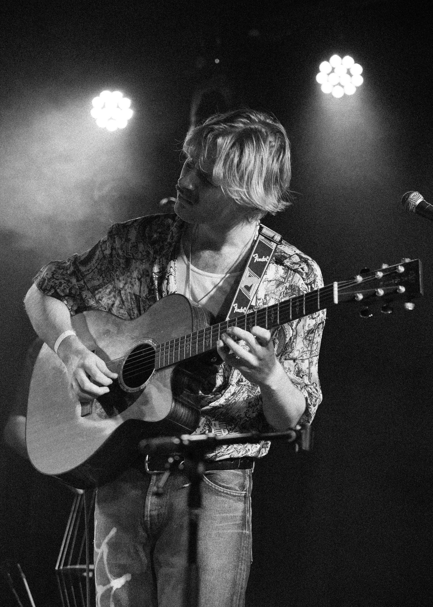 Young male musician playing acoustic guitar on stage under spotlights in black and white concert photo.