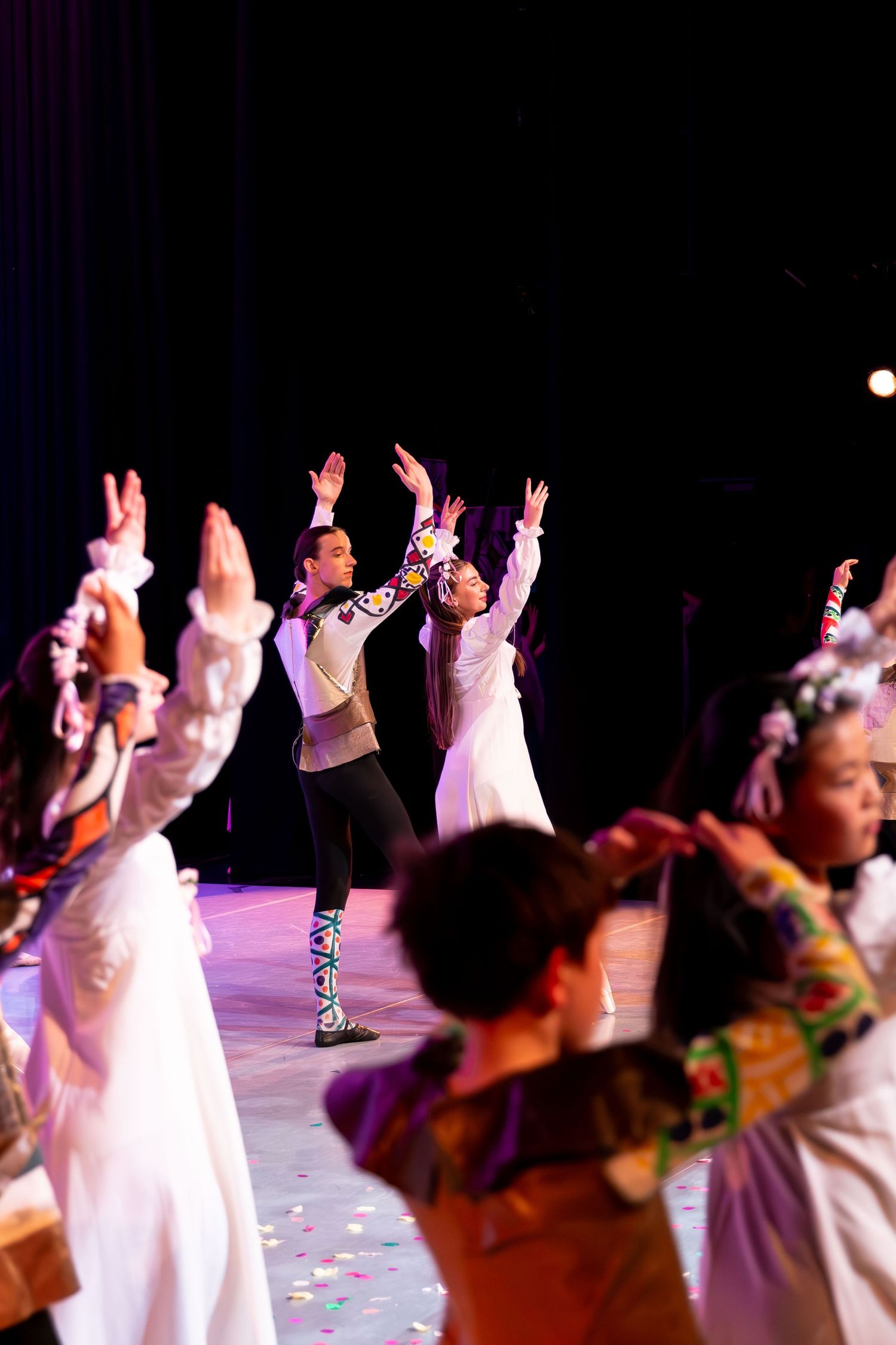Dancers in colorful traditional costumes perform on stage with arms raised under bright spotlights during a cultural dance show.