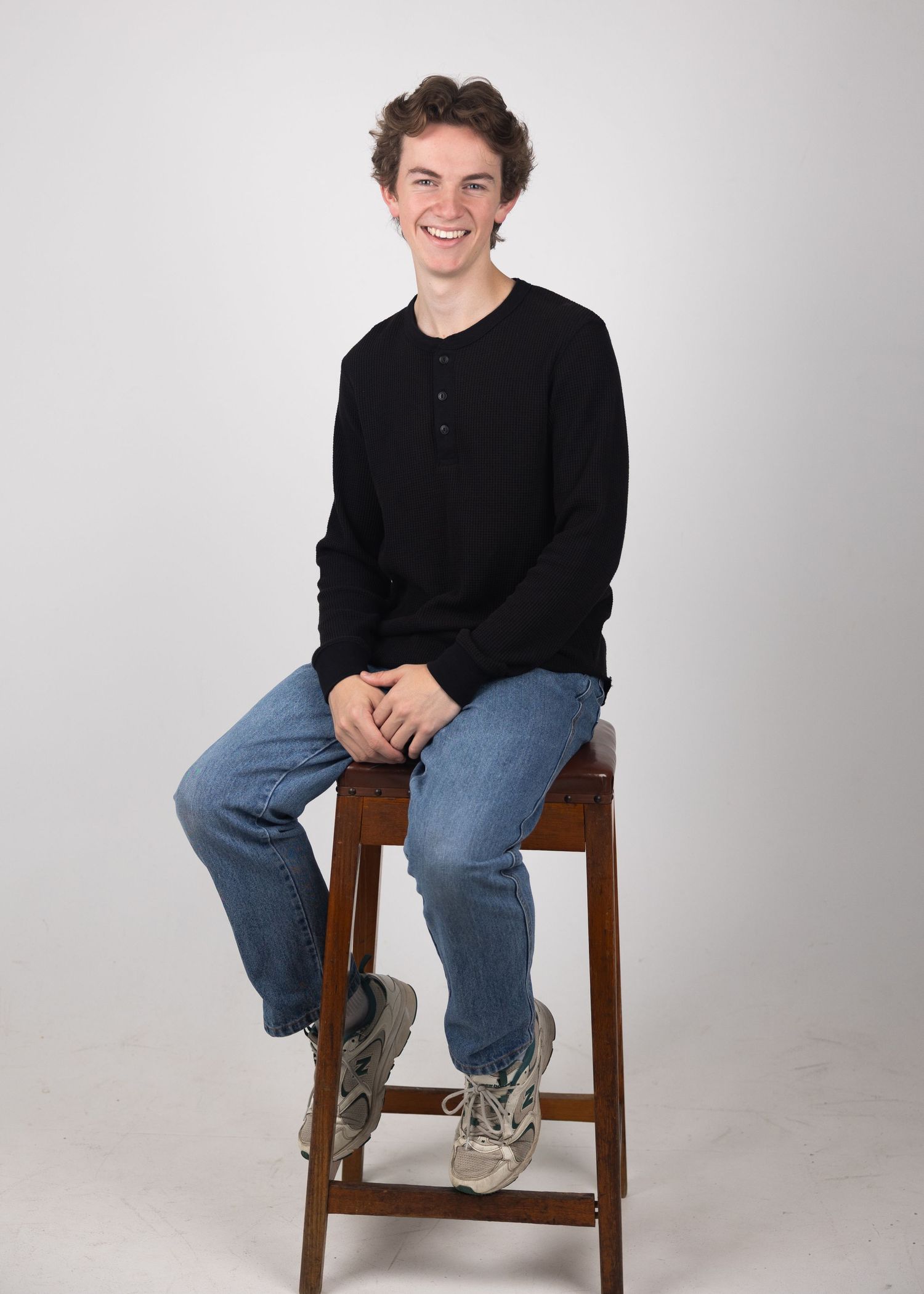 Young man in black henley and jeans sitting on a wooden stool, smiling against a white background.
