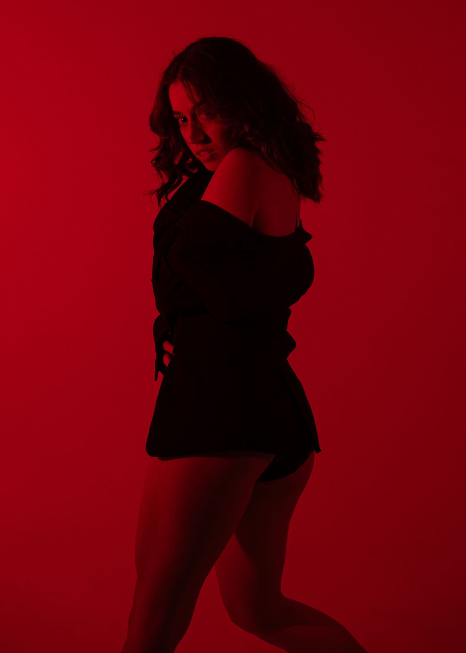 A woman in a black off-shoulder outfit poses against a vivid red background, looking back over her shoulder.