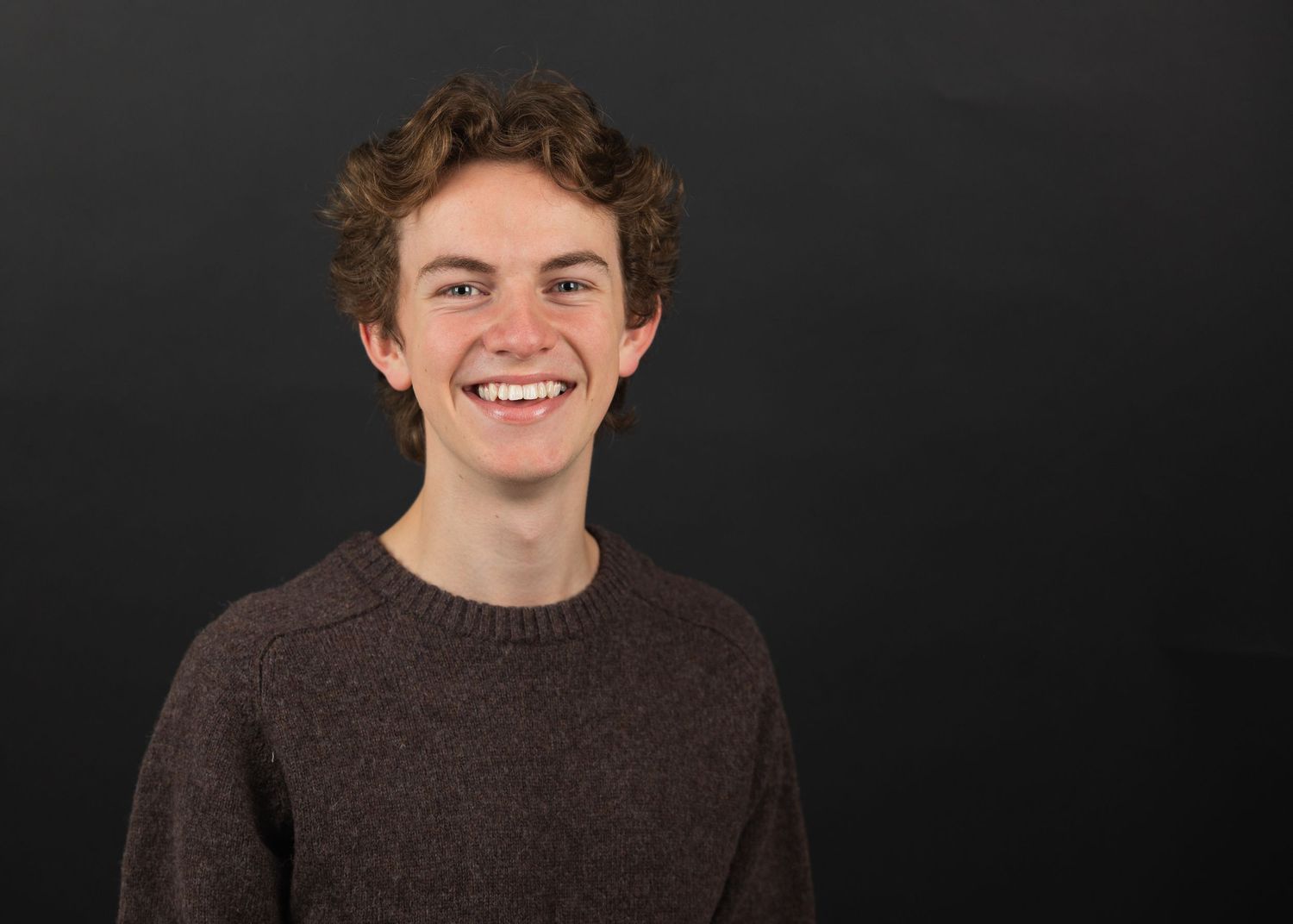 Smiling young man with curly hair wearing a brown knit sweater against a dark gray background.