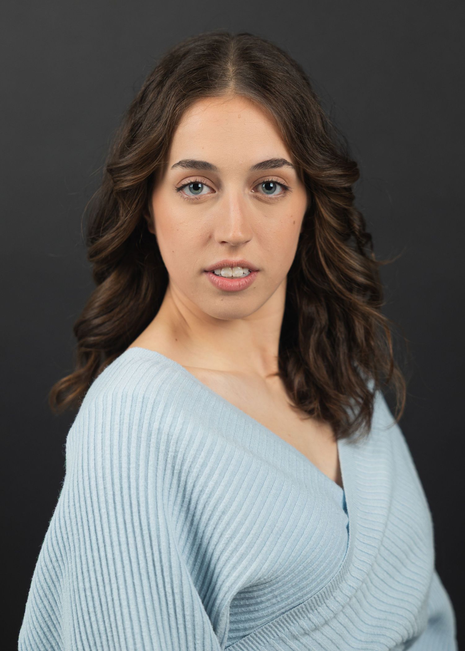 Young woman with wavy brown hair wearing a light blue ribbed sweater poses for a professional headshot photo.