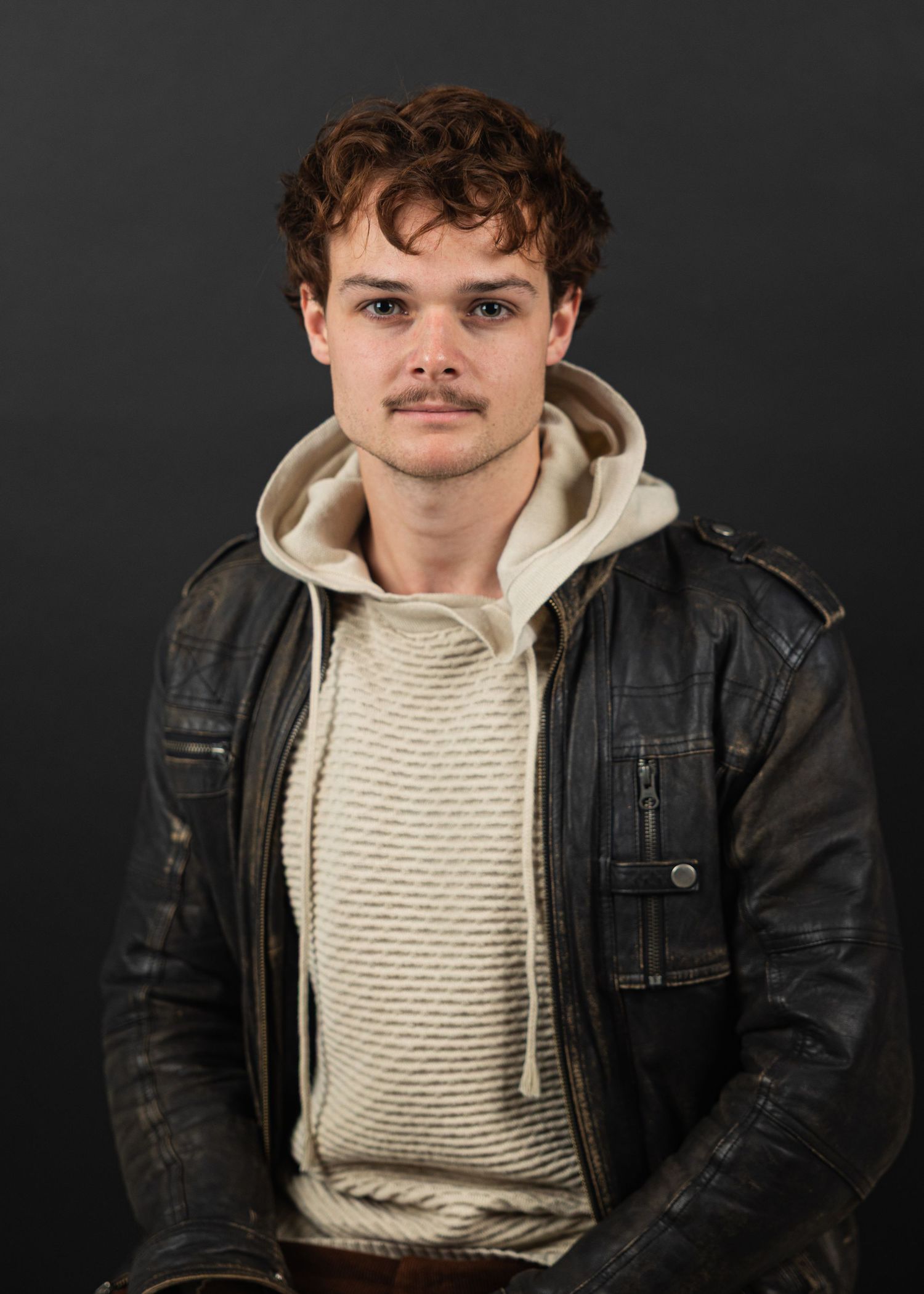 Young man with curly hair and mustache wearing a leather jacket and hoodie poses against a dark background.