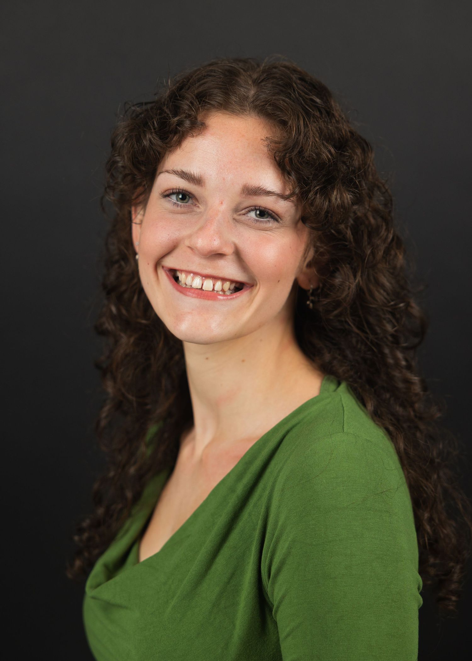 Smiling young woman with curly brown hair in a green top against a dark background.