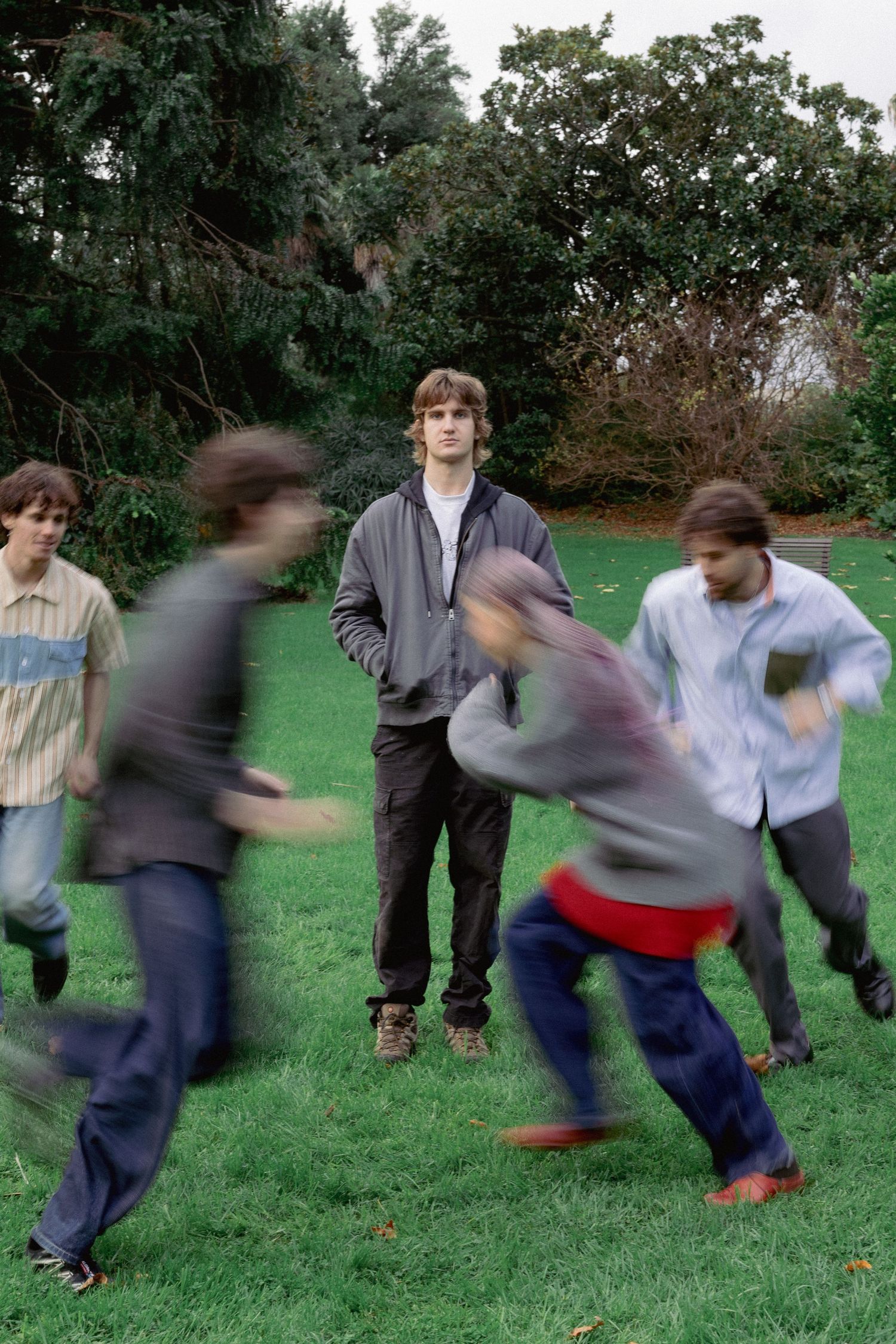 Five young men play in a park, with one standing still while others run around him in motion blur.