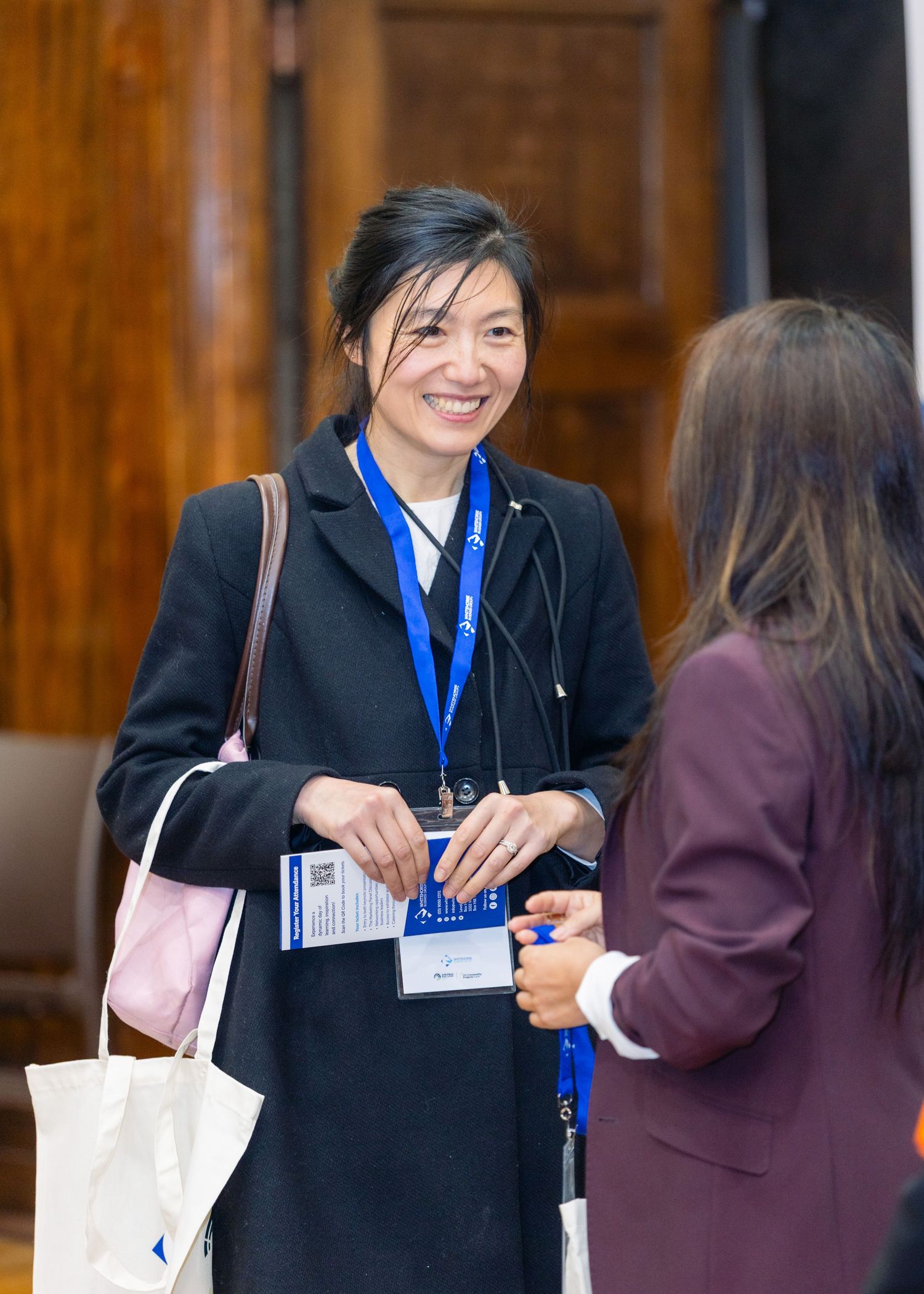 A smiling woman in a black coat with a blue lanyard badge converses with another attendee at a professional conference.