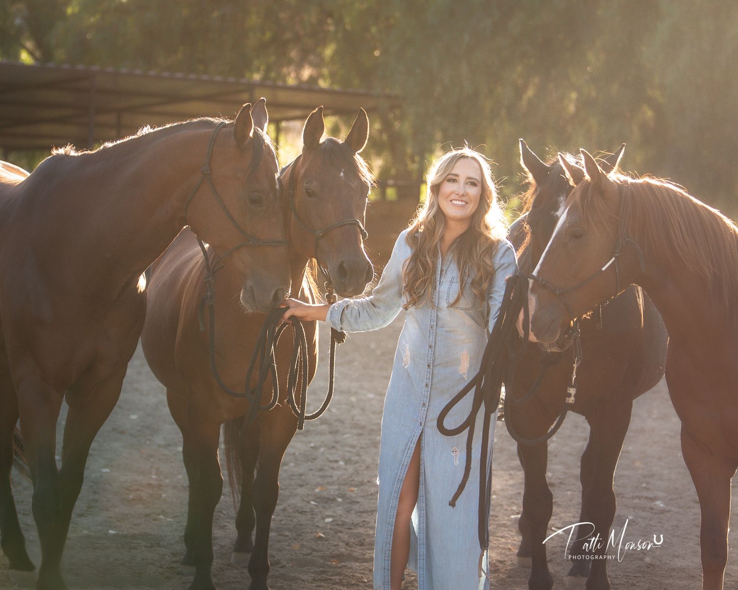 Woman in light blue dress stands gracefully between three chestnut horses in stunning golden sunset light.