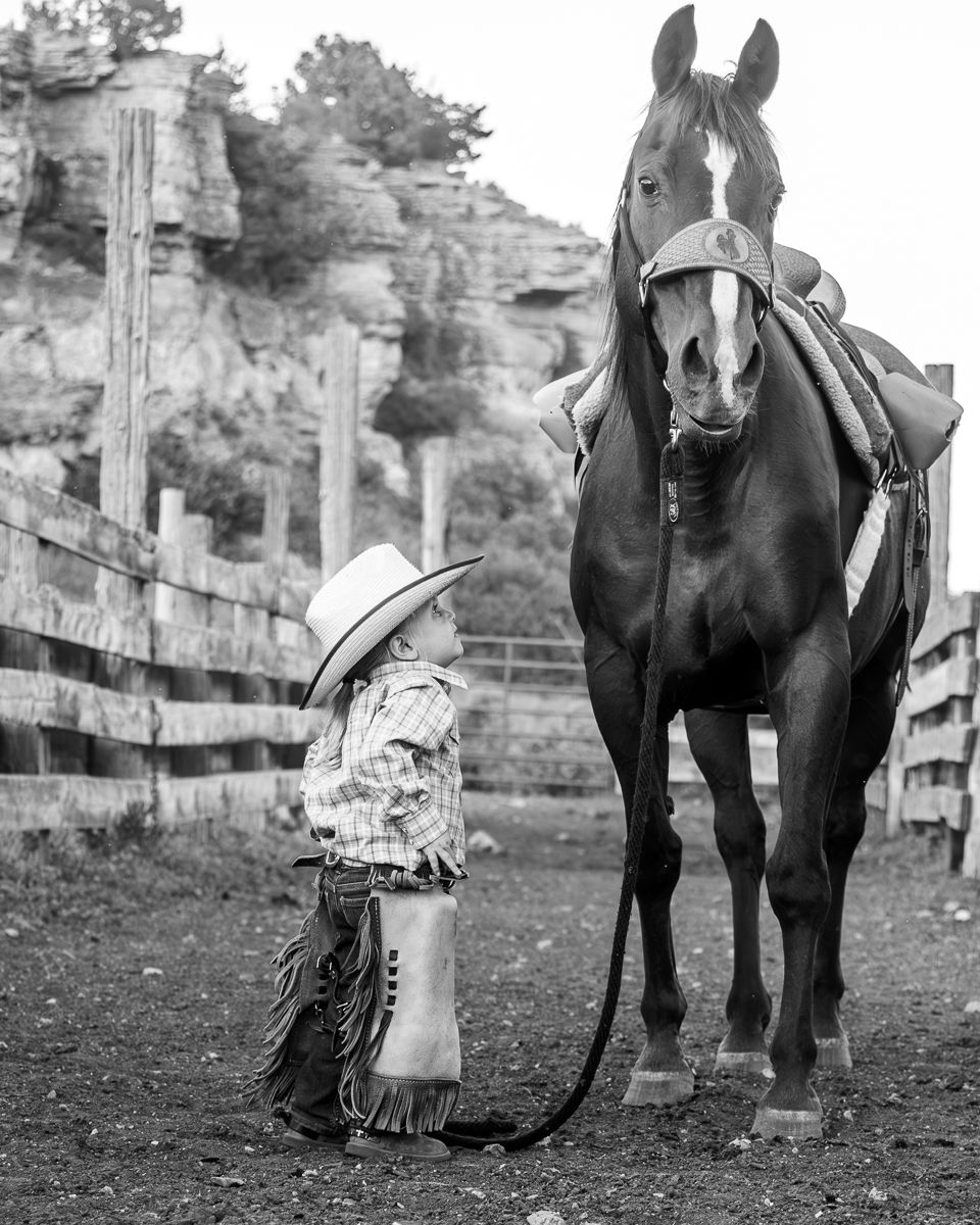 A small child in cowboy hat and chaps stands facing a saddled horse in a ranch corral, black and white photo.