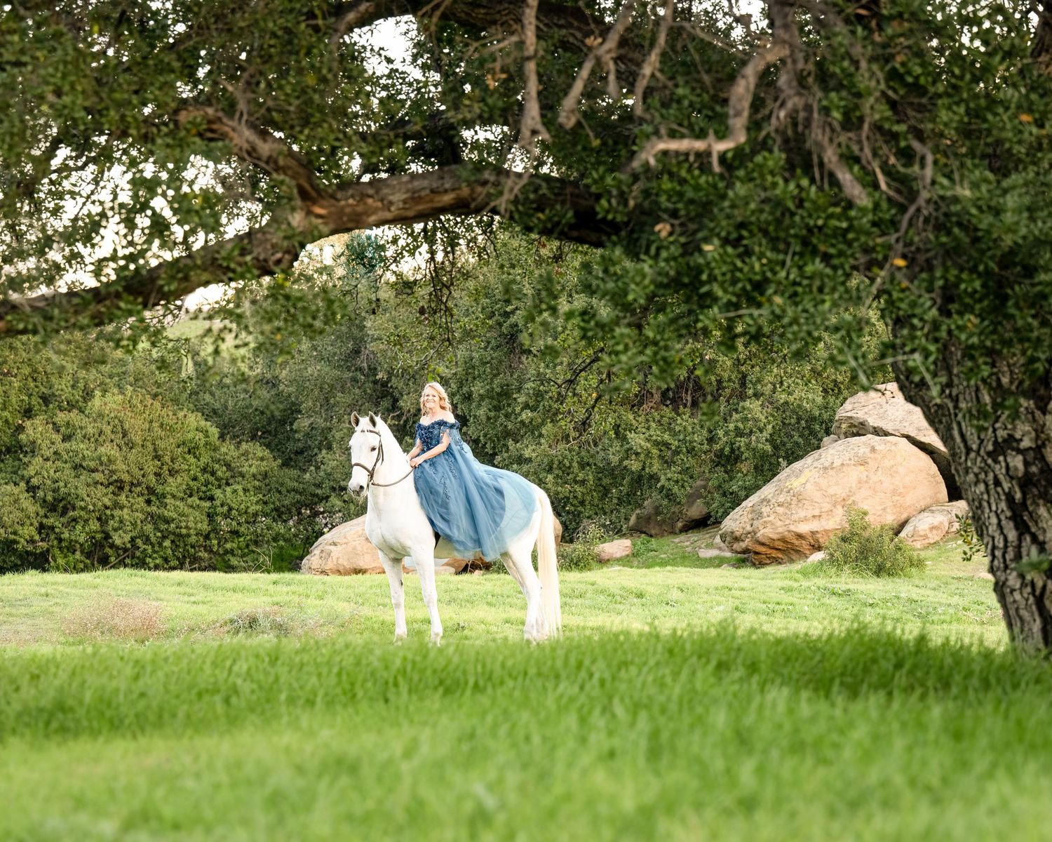 Women in a torques gown rides a white horse beneath a large oak tree in a lush green meadow in Southern California