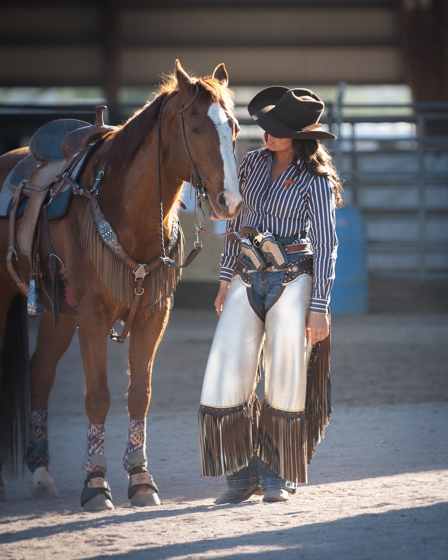 Cowgirl in fringed chaps and black hat stands beside a chestnut horse in an indoor riding arena.