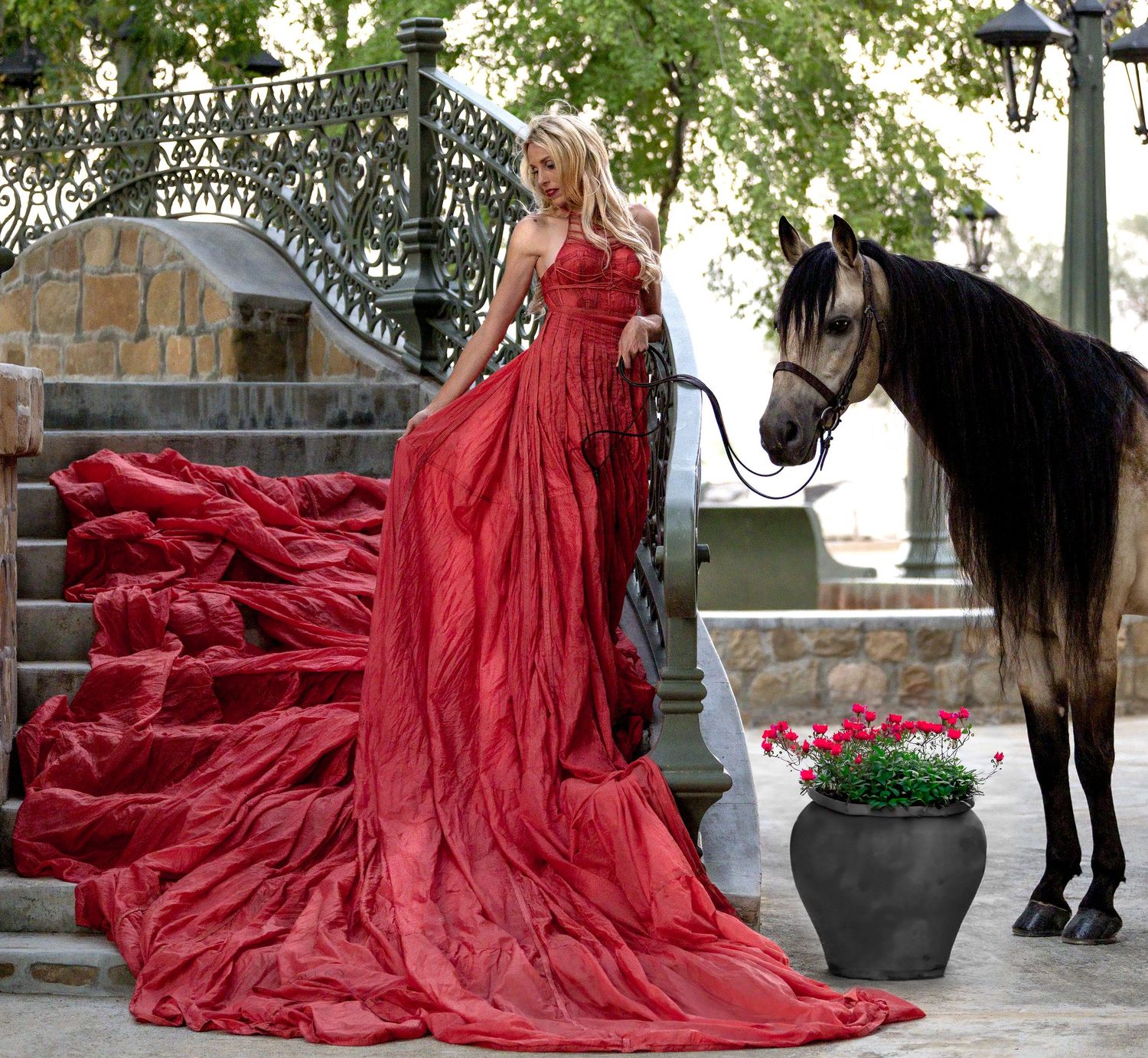 Blonde woman in elegant red gown with long train stands beside a dark horse on ornate outdoor staircase.