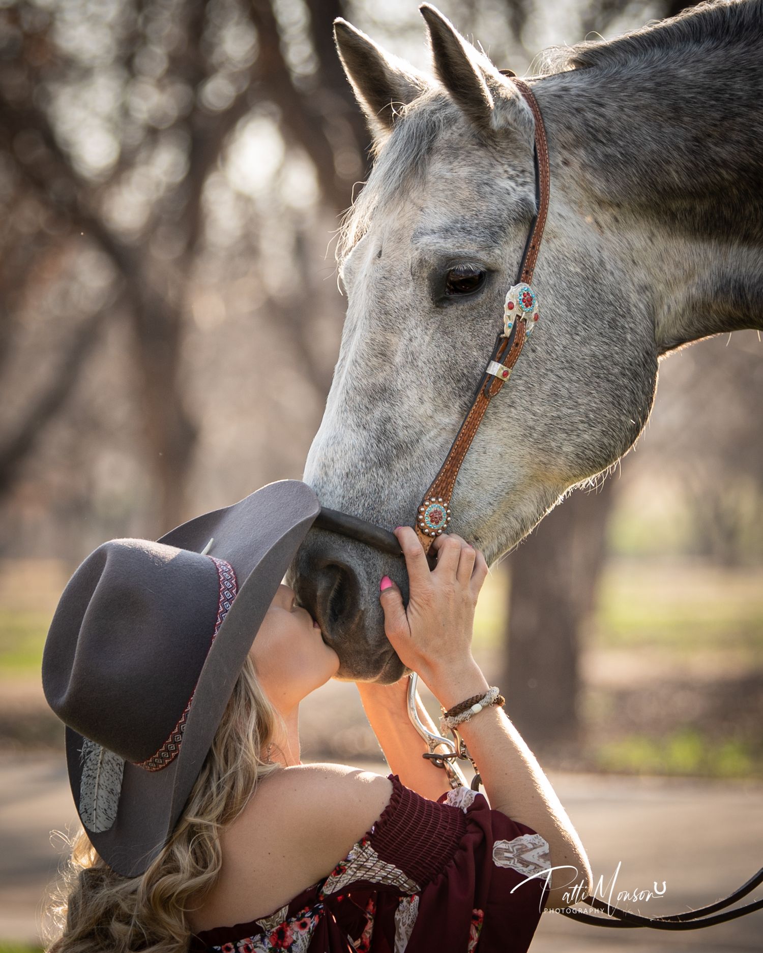 Blonde woman in cowboy hat gently kissing the nose of a dapple grey horse outdoors.