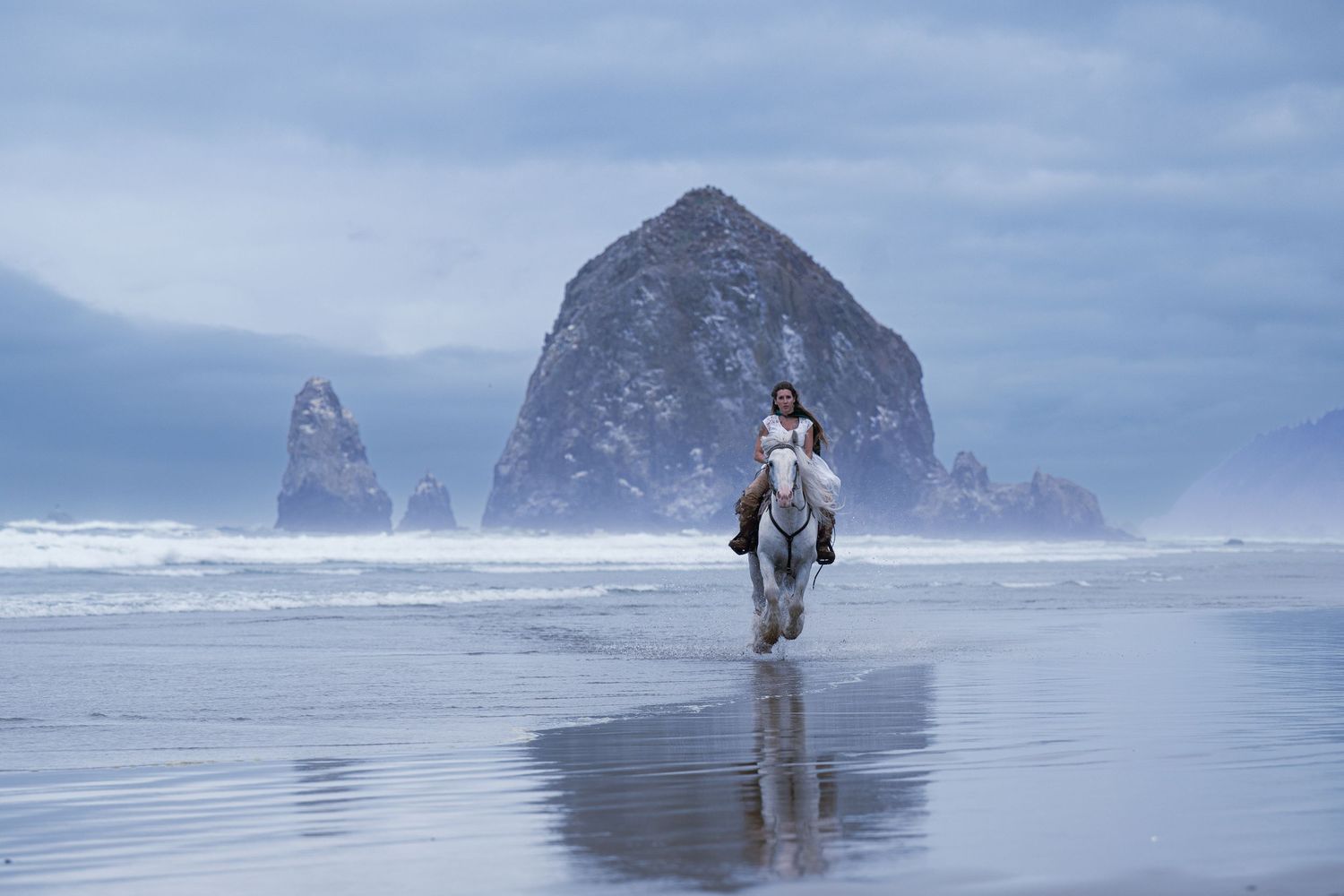 Rider gallops a white horse along a misty beach with Haystack Rock visible in the background.