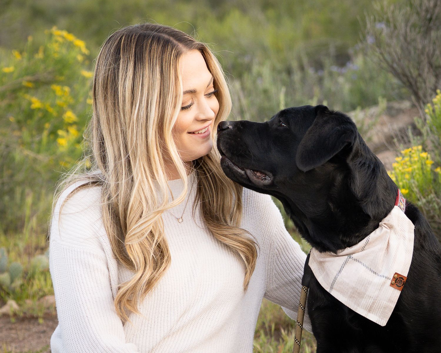 Blonde woman smiling as black Labrador dog licks her face in a desert garden setting.