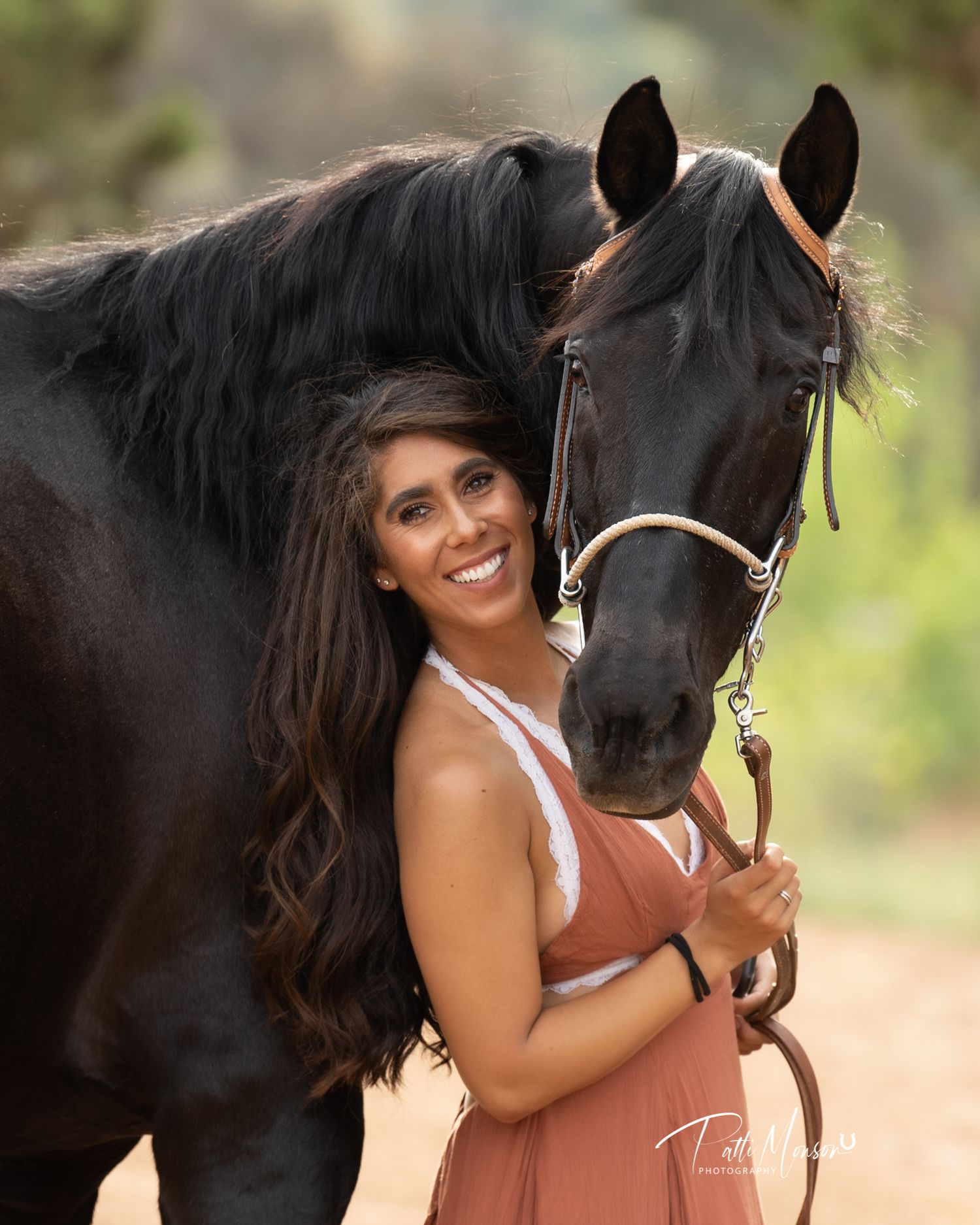Smiling brunette woman in pink dress embracing black Friesian horse outdoors.