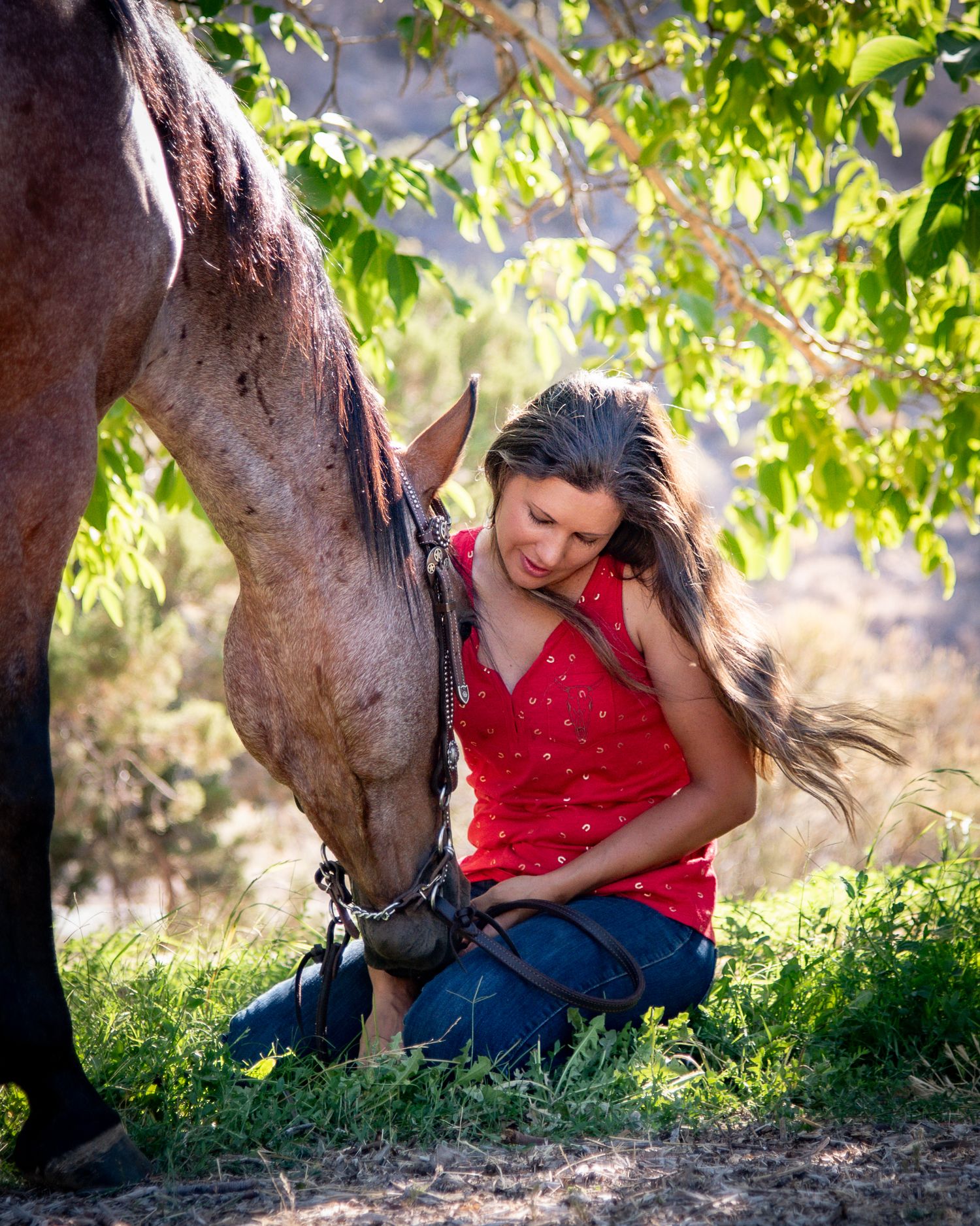 Young woman in red top sits on grass bonding with a brown horse under green trees in natural light.