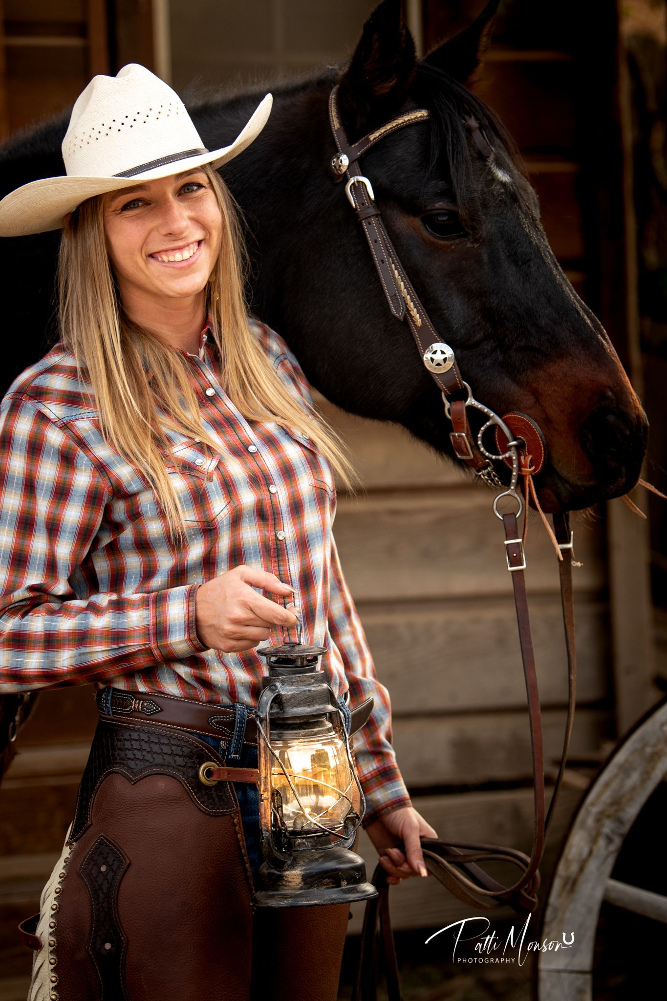 Smiling cowgirl in plaid shirt and white hat holding vintage lantern beside black horse.